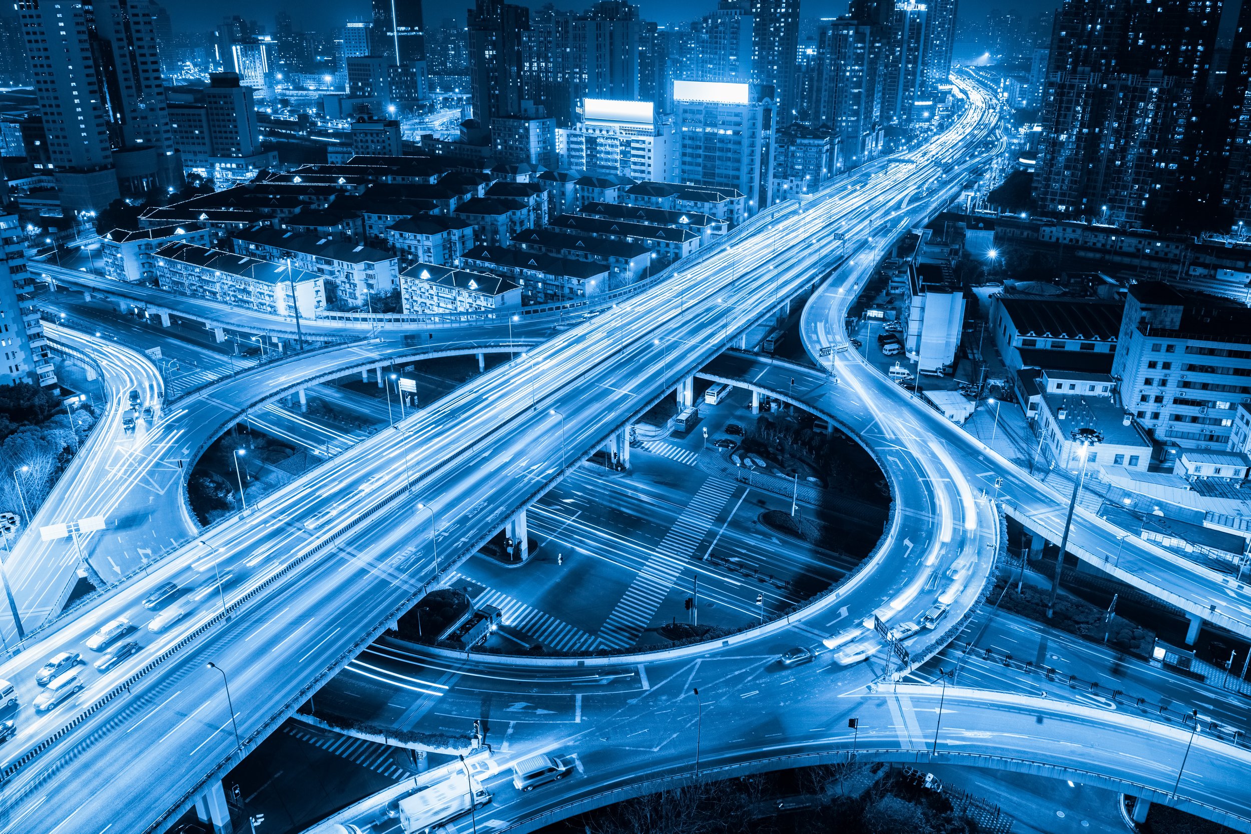 A cityscape at night with illuminated high-rise buildings and busy highways, seen from above with streaks of vehicle lights on the roads.