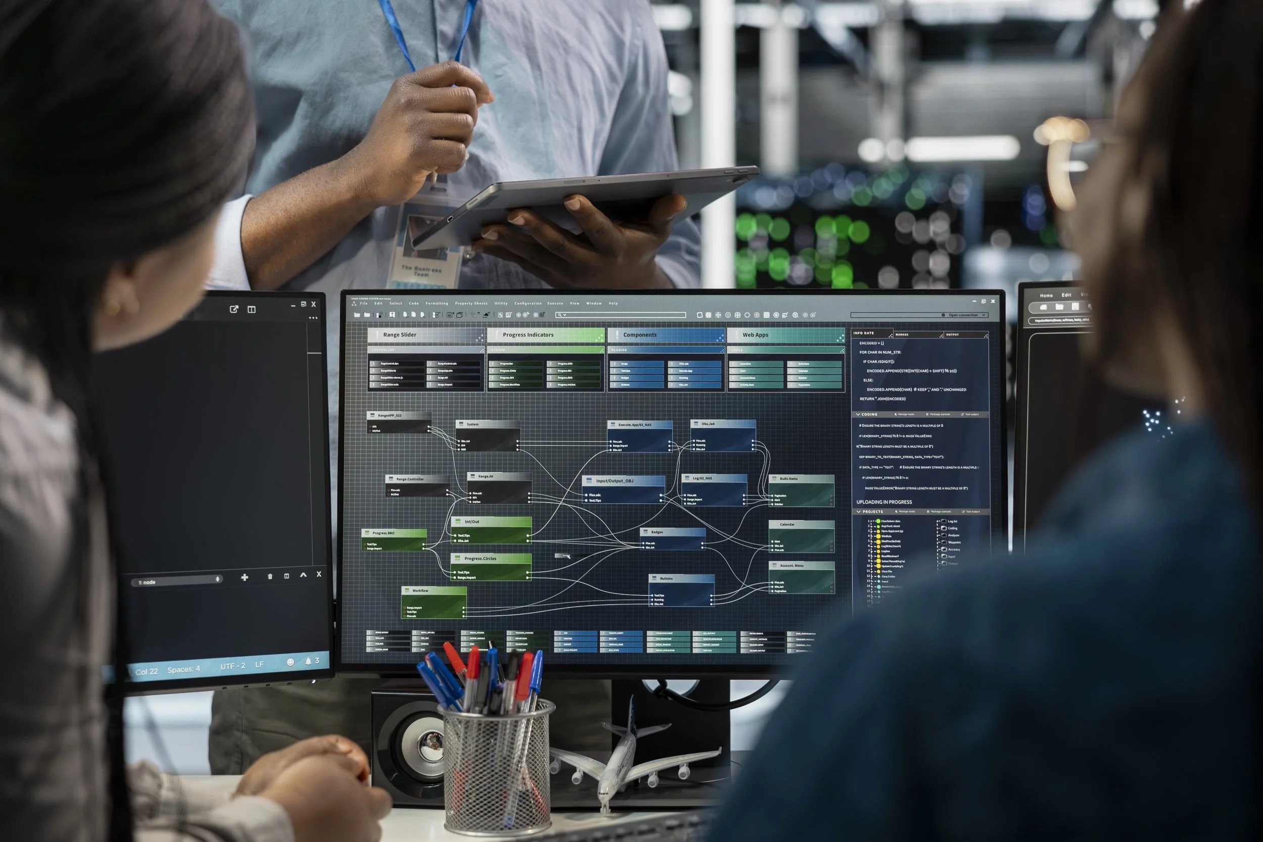 People working on a computer screen displaying a complex flowchart or mind map with multiple colored nodes and text, with a person standing nearby looking at a tablet and others seated at the desk.