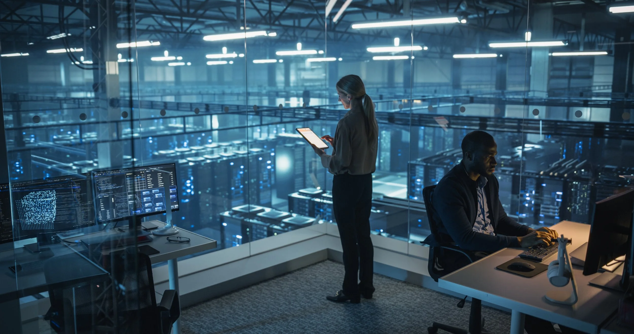 Two professionals working in a high-tech data center or server room with multiple computer monitors; a woman stands and looks at a tablet, a man sits at a desk typing on a keyboard, with glass walls and server racks visible in the background.