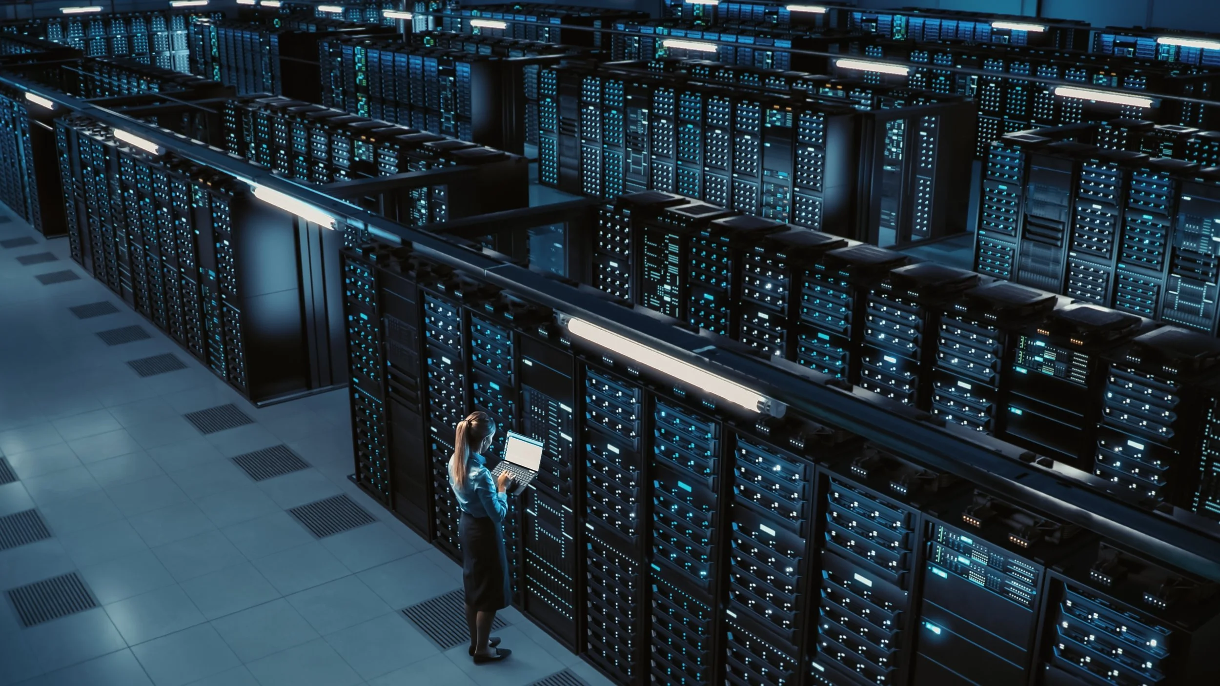 A woman in a server room using a laptop, surrounded by tall server racks with blue and green lights.