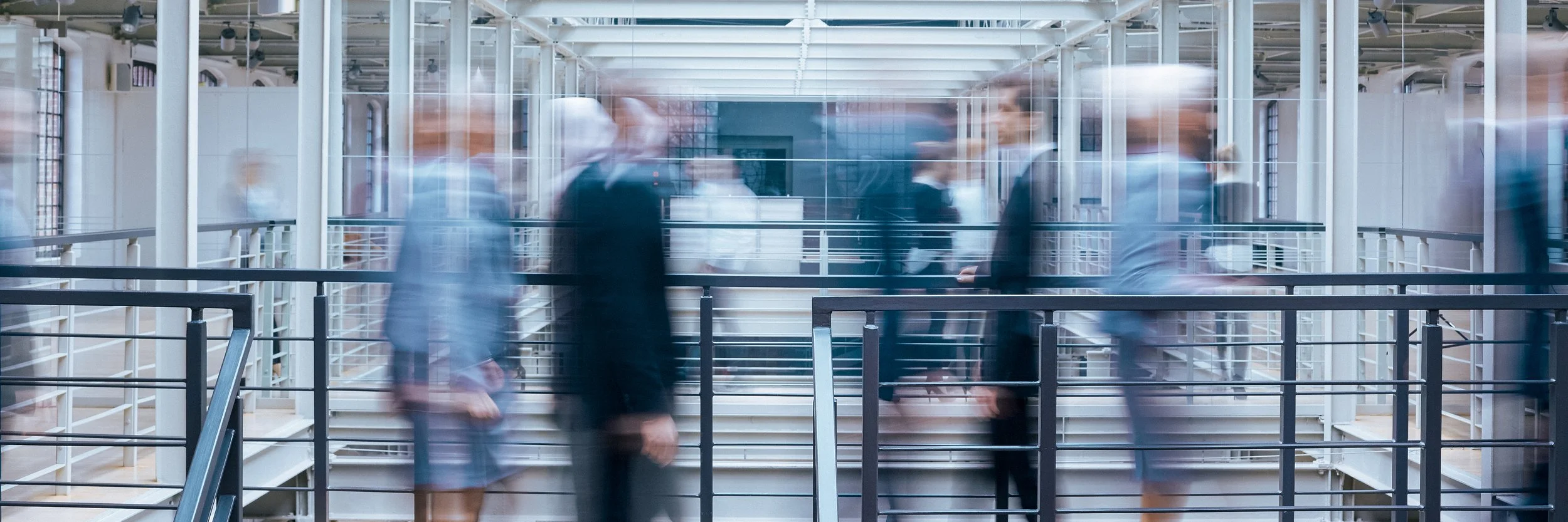 Blurred motion of business professionals walking inside a modern office building with glass walls and metal railings.