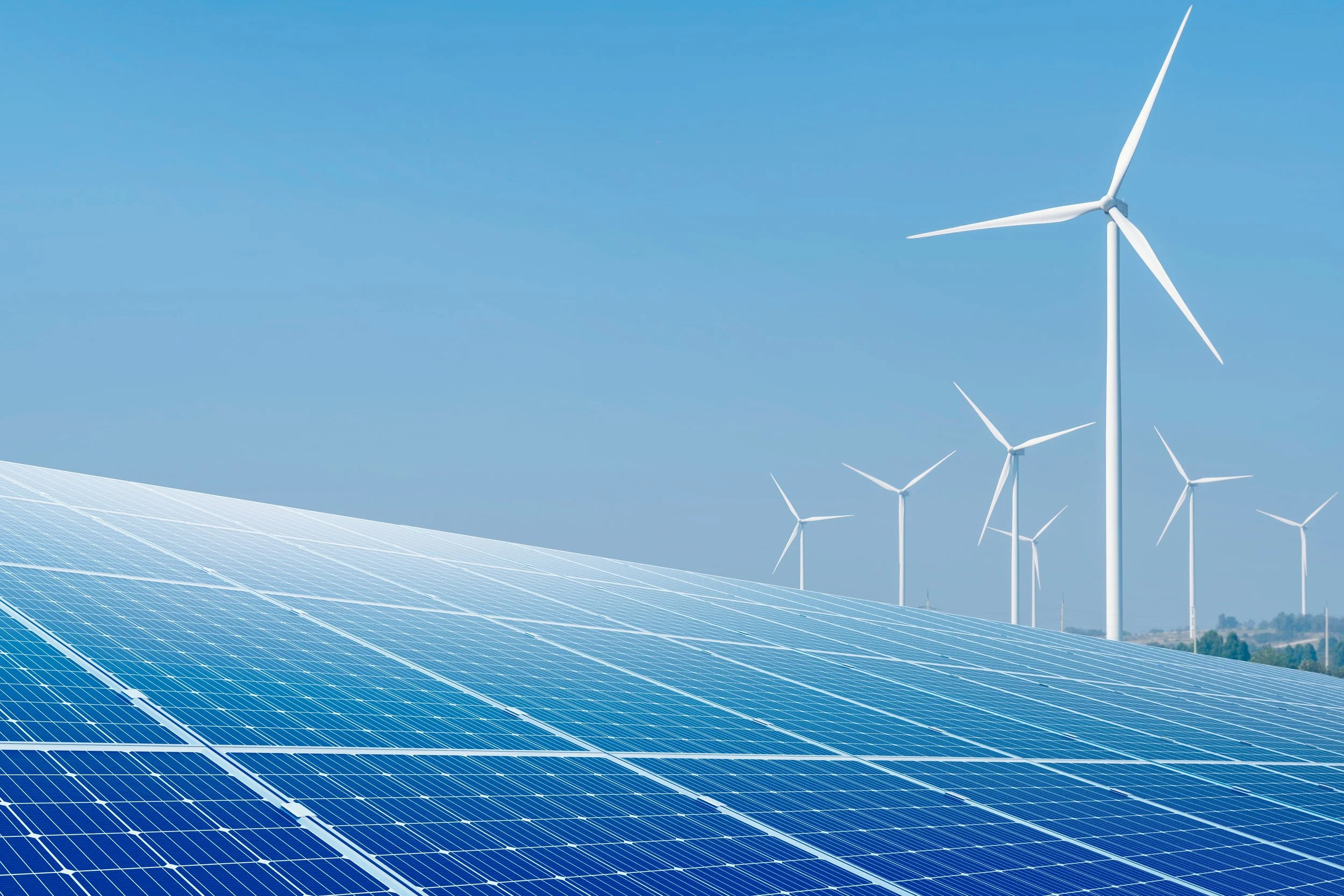 A large solar panel array in the foreground with a wind turbine farm in the background, set against a clear blue sky.