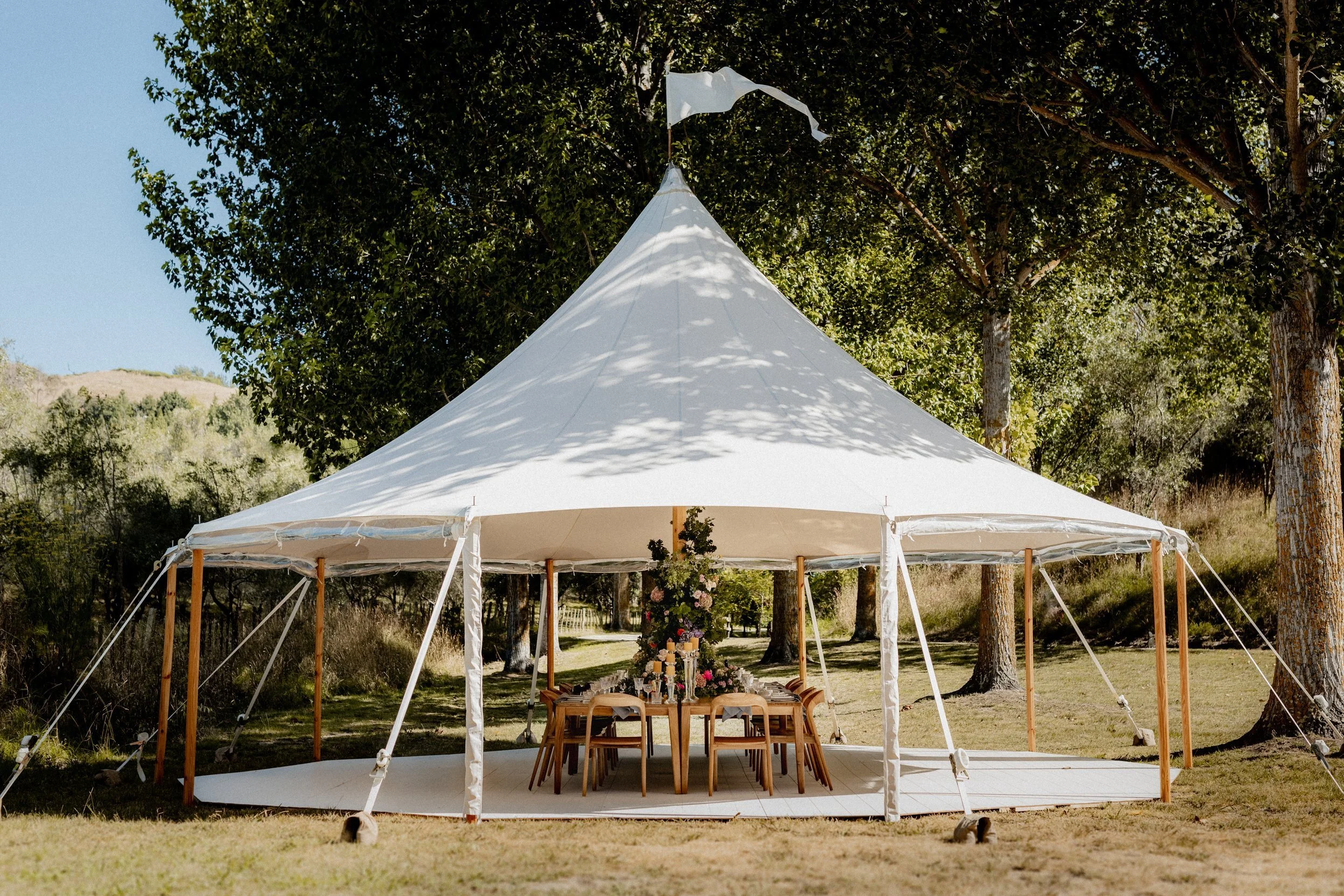 A white outdoor event tent set up in a grassy area with large trees, decorated with a central floral arrangement and candles, likely for a celebration or gathering.