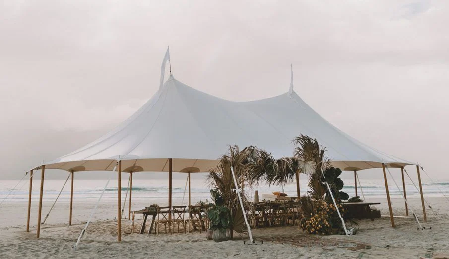 Large white event tent set up on sandy beach with tables, chairs, and tropical plants underneath, with the ocean in the background.