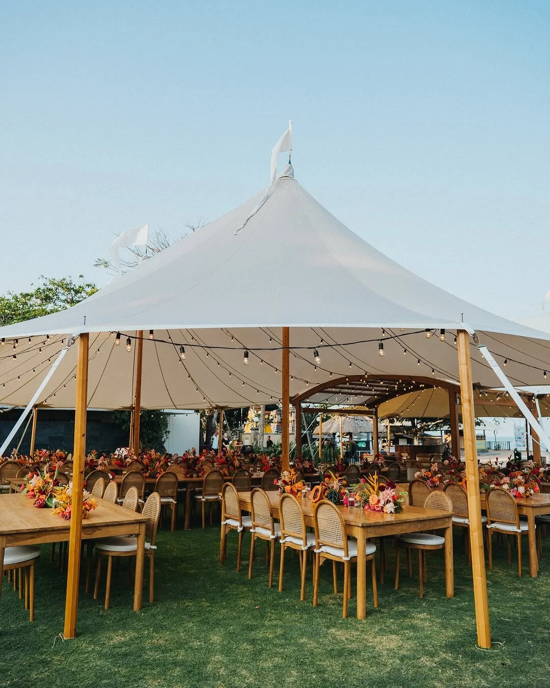 Outdoor event setup with a large white tent, tables and chairs, adorned with colorful floral arrangements, set on grass.