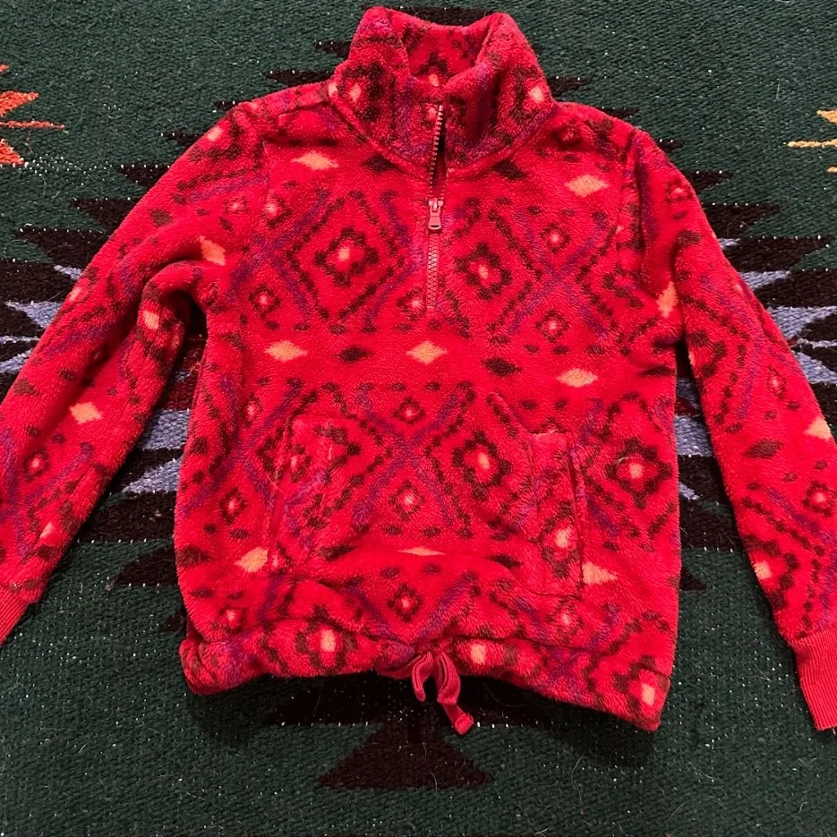 A red fleece pullover with a black geometric pattern, a half-zip collar, and a drawstring hem, lying on a dark green woven textile background.