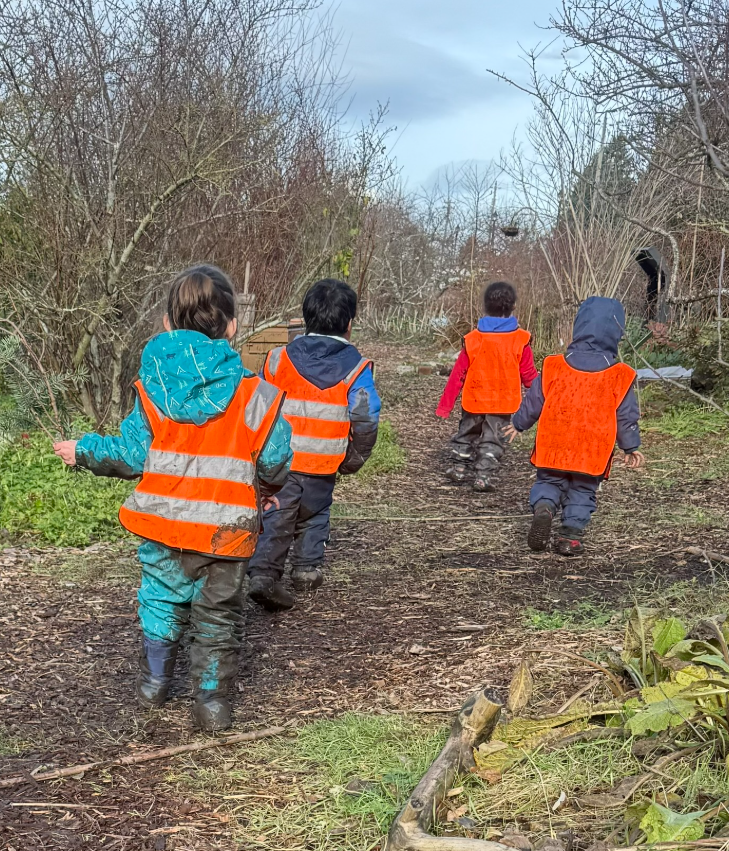 Two children wearing yellow safety vests playing balancing on a large rock outdoors in a wooded area.