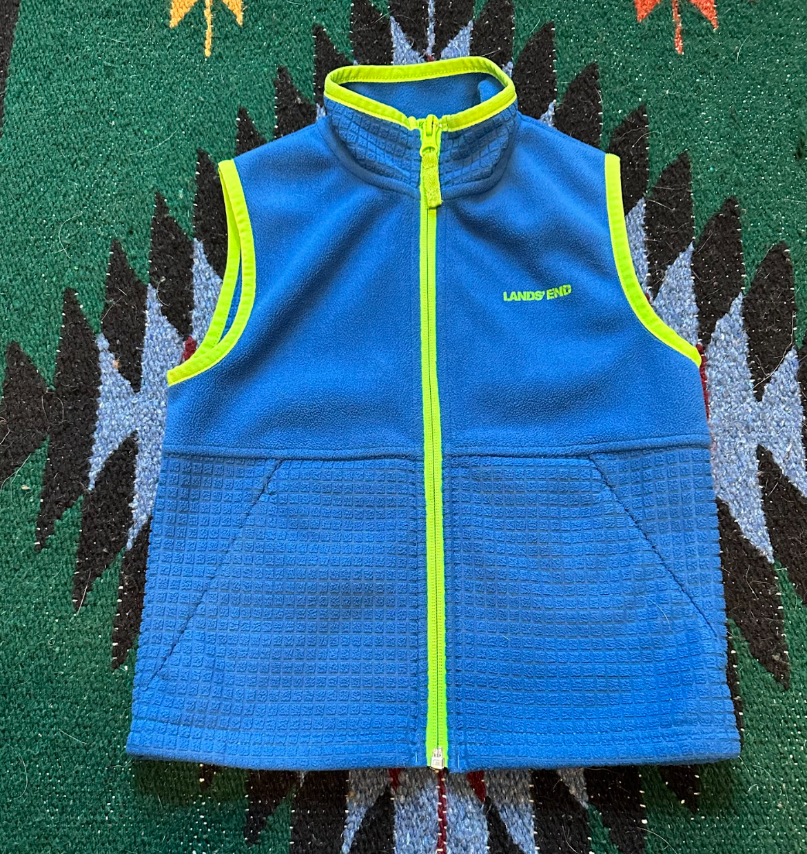 Blue sleeveless fleece vest with neon green accents and zipper, and the word 'LAND'S END' embroidered on the chest, lying on a patterned green carpet.