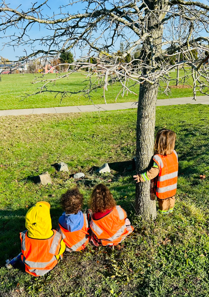 Child holding a blue bucket filled with muddy water, using a wooden stick to stir, outdoors on a muddy ground with small plants and stones.