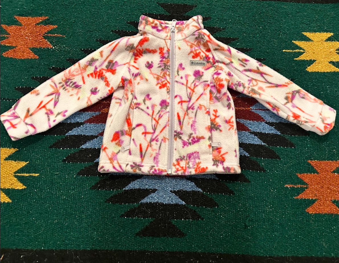 A children's fleece jacket with a floral pattern in pink, purple, and red on a white background, lying on a colorful geometric patterned textile.