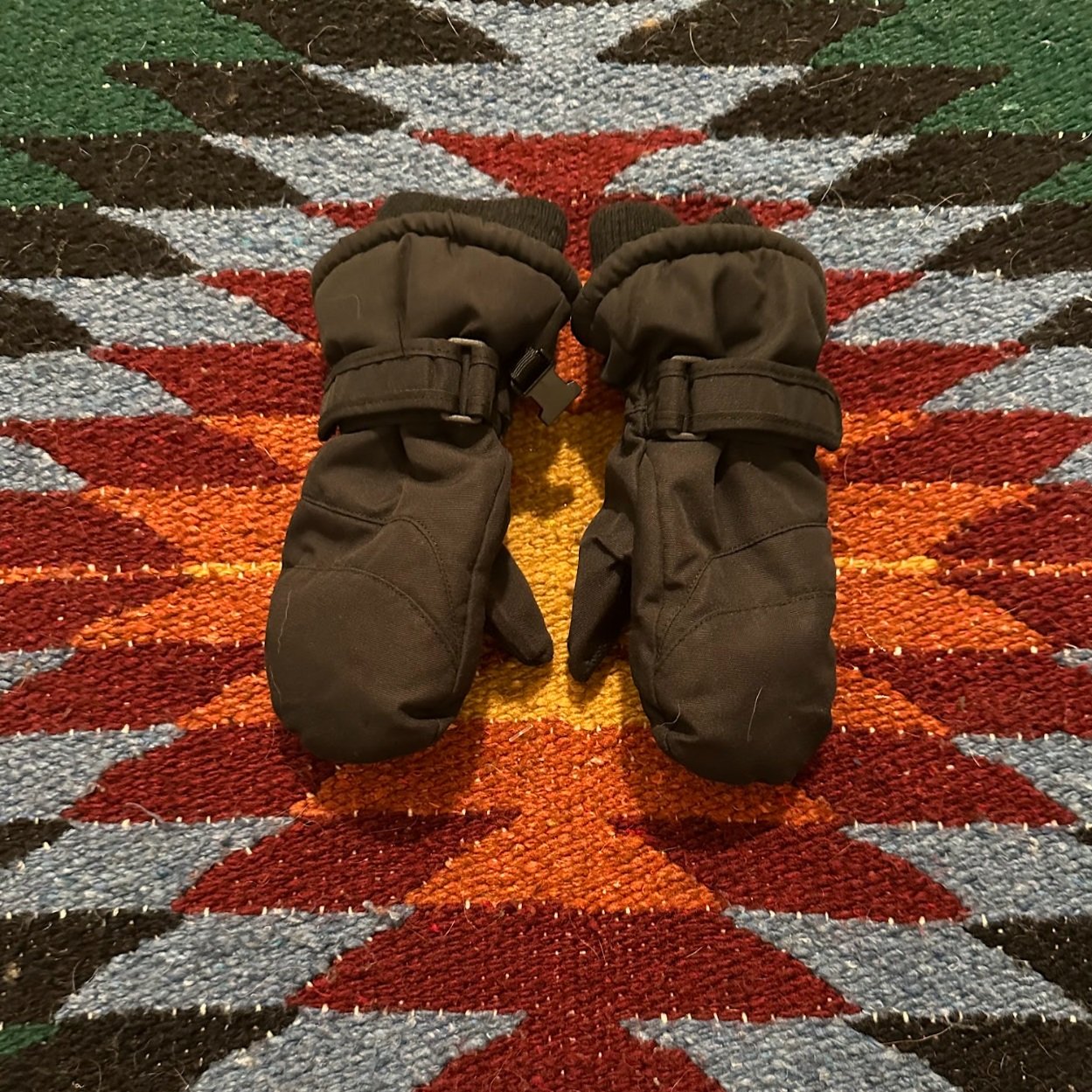 Pair of black winter mittens placed on a colorful geometric patterned rug.
