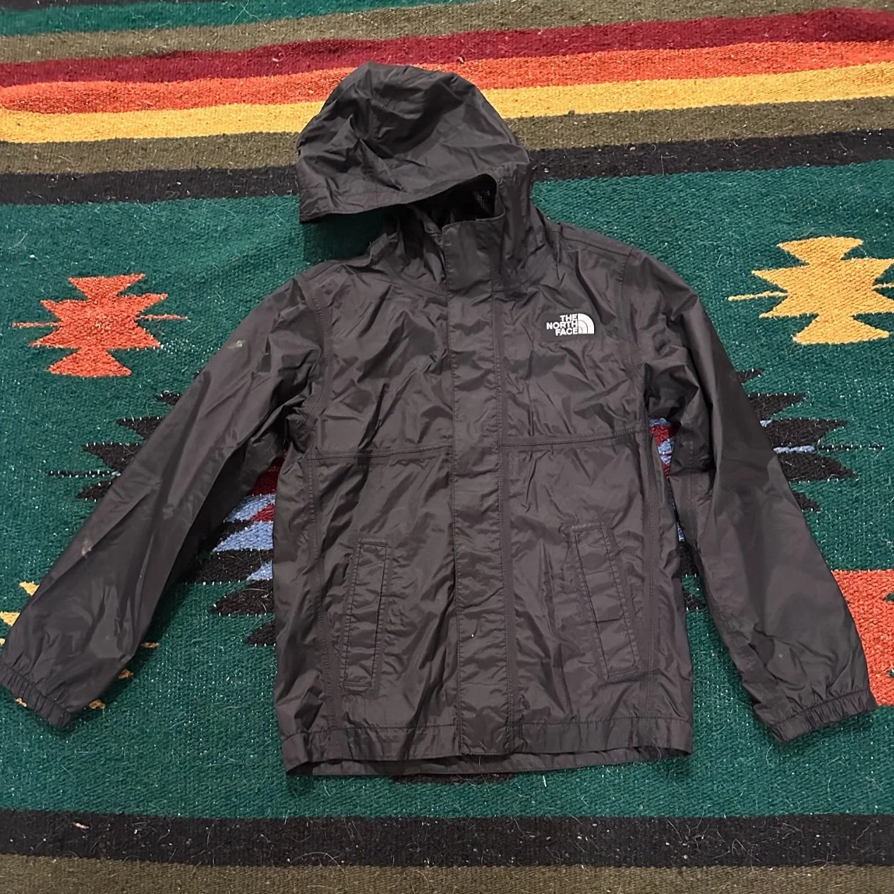 Black North Face waterproof jacket with hood laid flat on a colorful patterned rug.