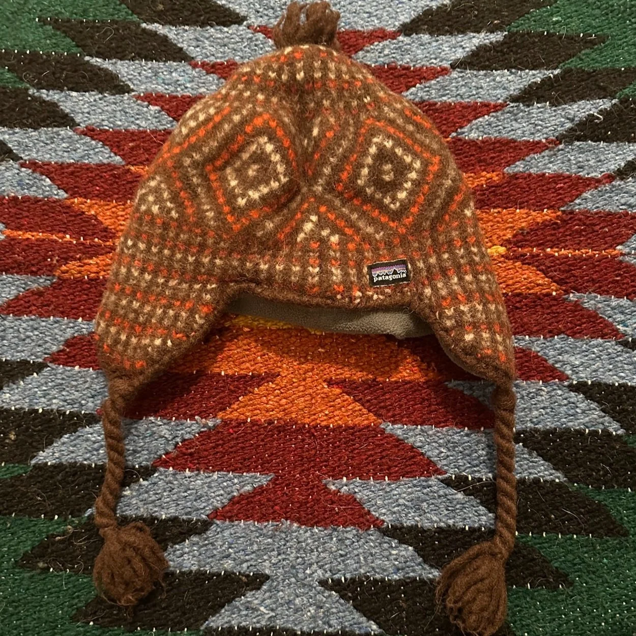 A brown, patterned Patagonia winter hat with ear flaps and braided tassels, placed on a colorful, geometric patterned rug.