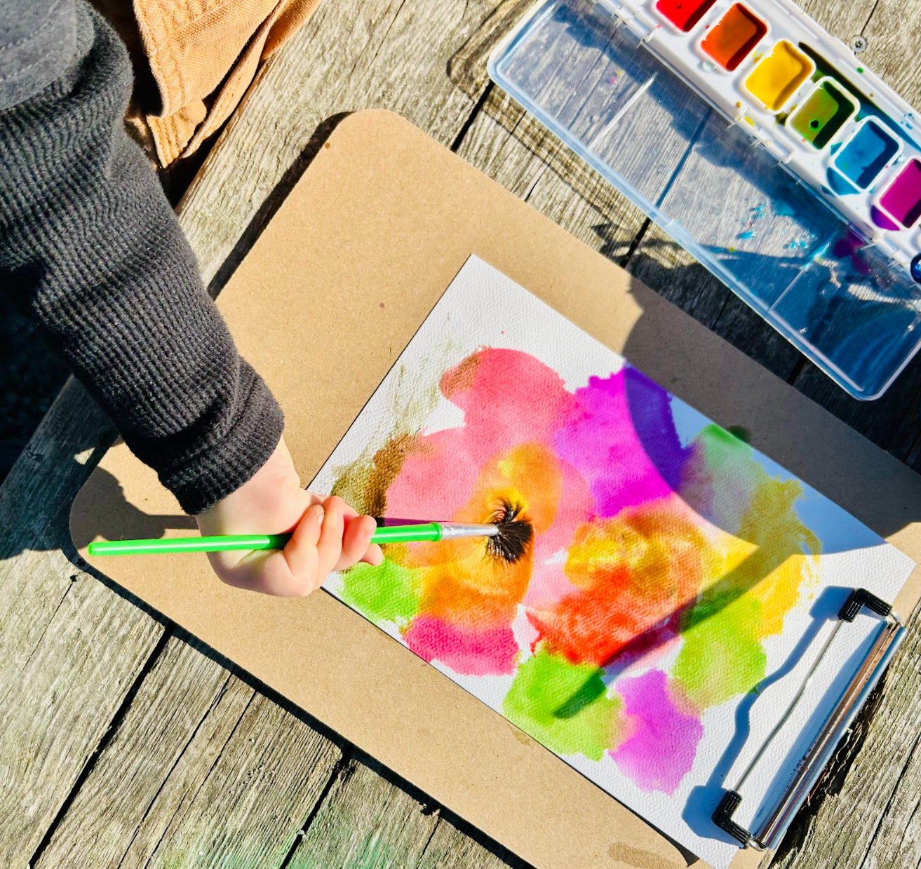 Young child with messy brown hair wearing a yellow raincoat and blue rain boots, sitting on a multicolored blanket outdoors, examining purple paint on her hands.