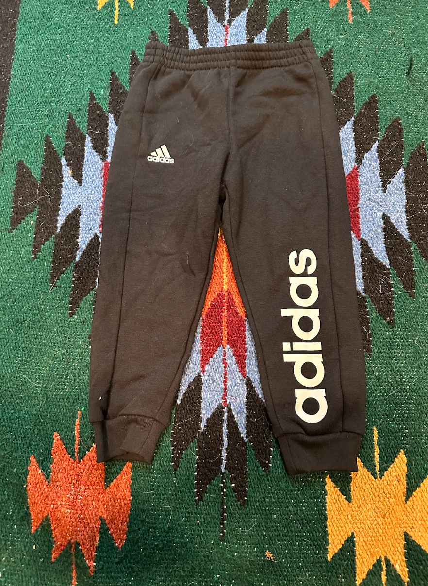 Black Adidas sweatpants with the Adidas logo on the left thigh and the brand name printed vertically on the right leg, laid flat on a colorful, patterned rug.