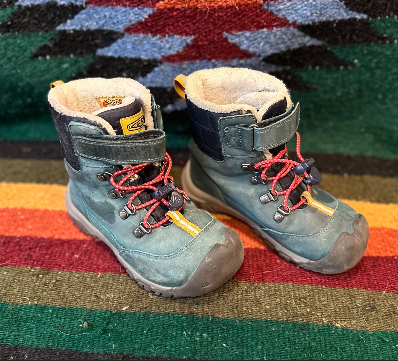Pair of green and black hiking boots with red laces on a colorful striped rug with a flower pattern in the background.