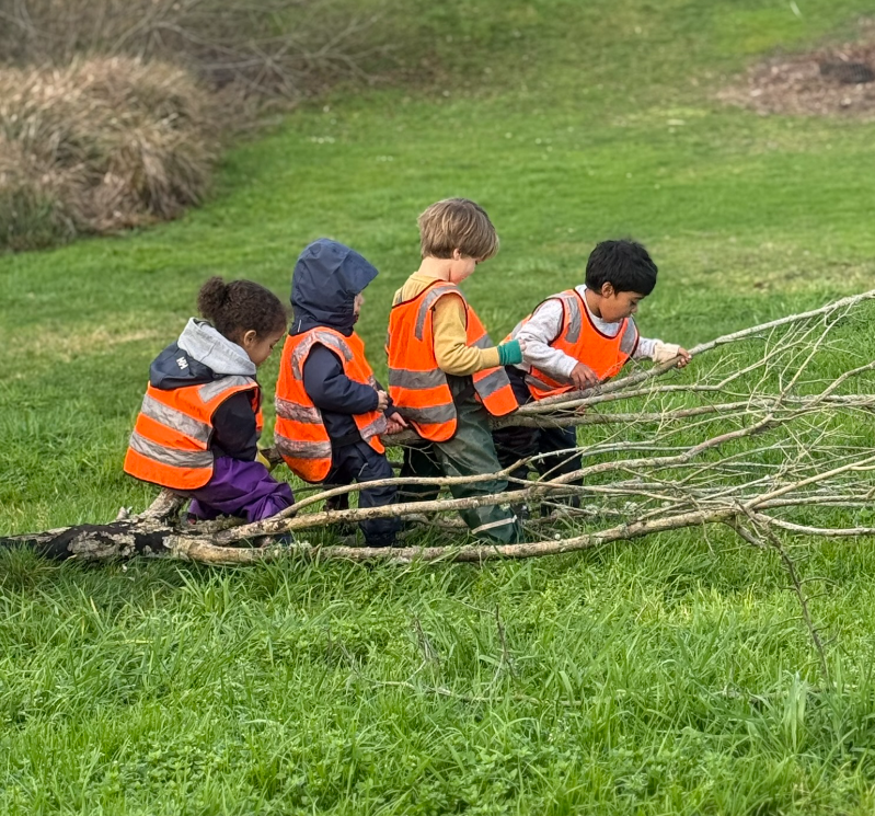 A smiling young girl with muddy face and hair, wearing a yellow rain jacket and a neon yellow safety vest, playing outdoors near a play structure with wood chips on a sunny day.