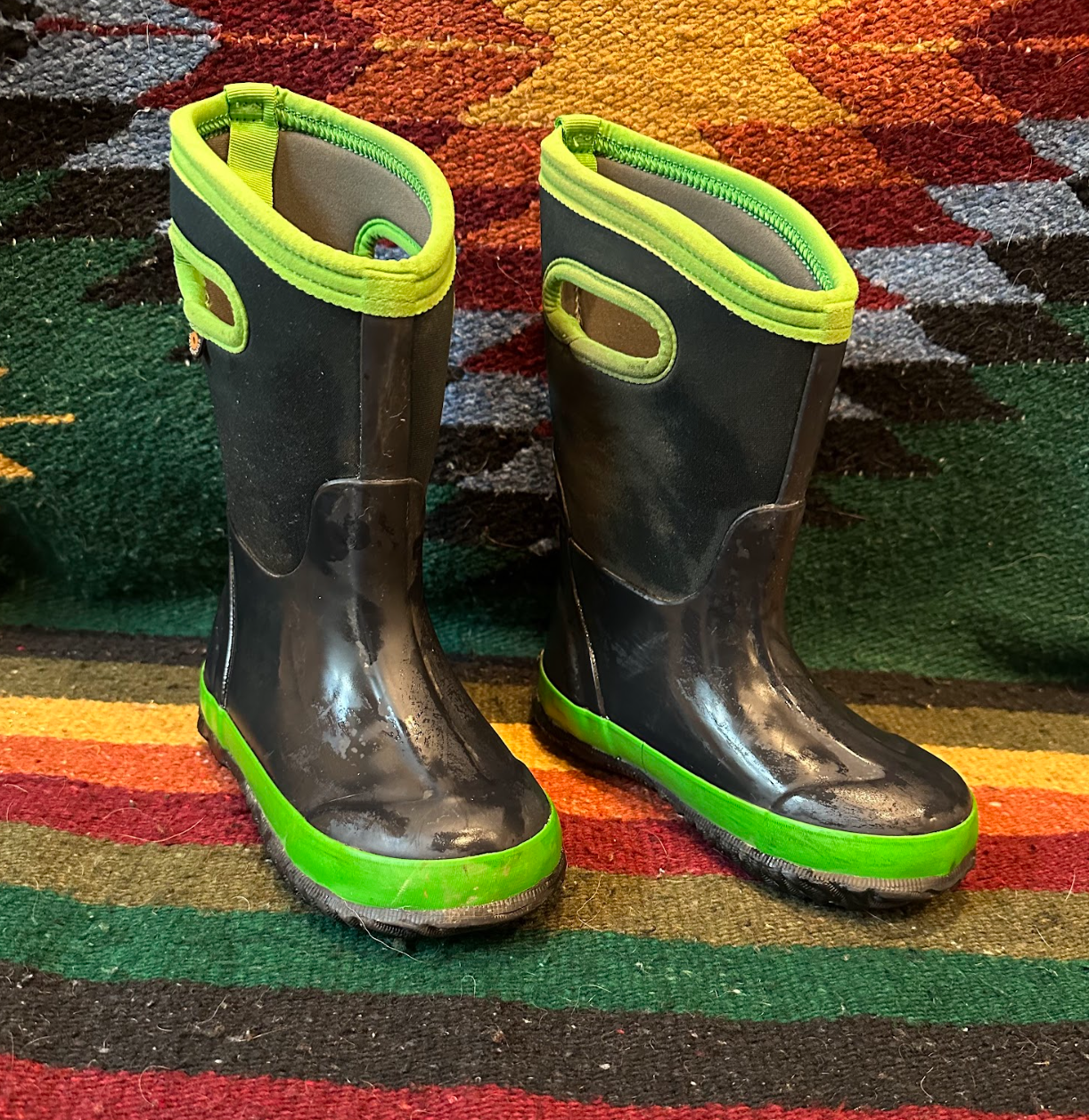 Pair of black rubber rain boots with neon green accents, including the soles and top trim, positioned on a multicolored patterned carpet.