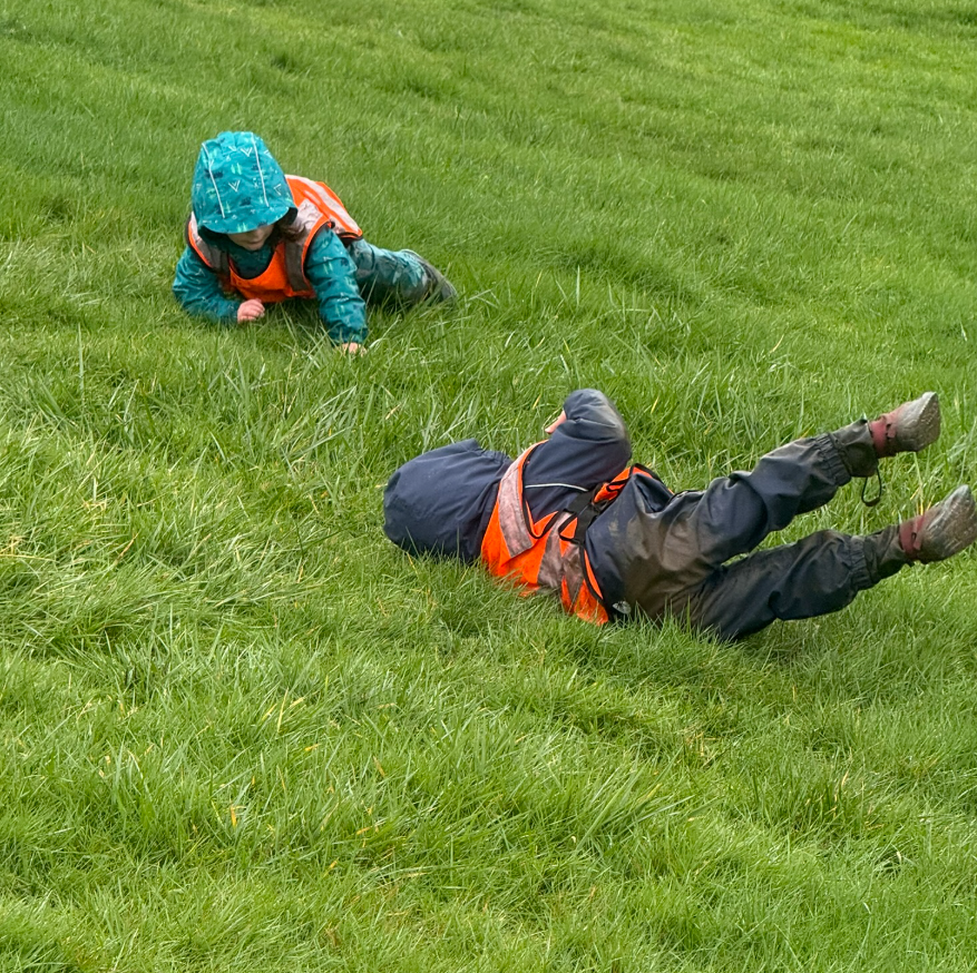 Three children walking on a dirt patch in a park, wearing colorful capes, with a city skyline, including the Space Needle, in the background.