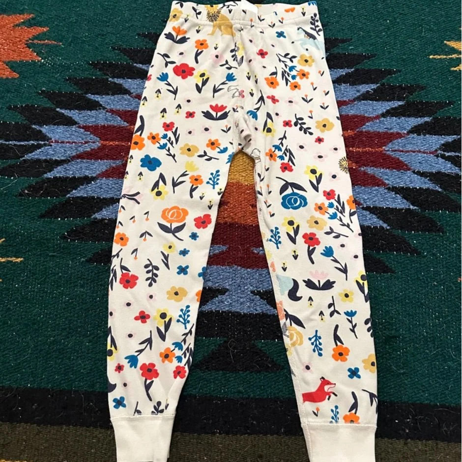 Pair of white pants with colorful floral patterns and small animal illustrations, laid on a patterned rug.