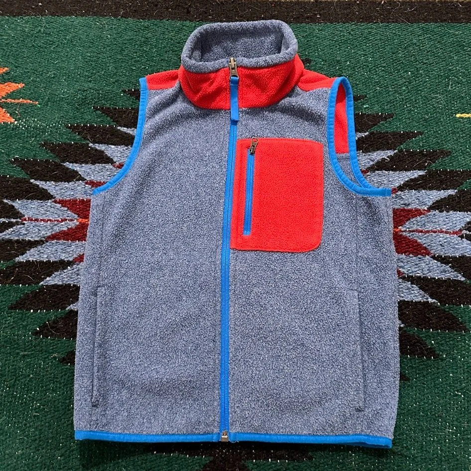 Gray sleeveless fleece vest with red accents, blue zipper, and chest pocket, laid flat on a multicolored patterned rug.