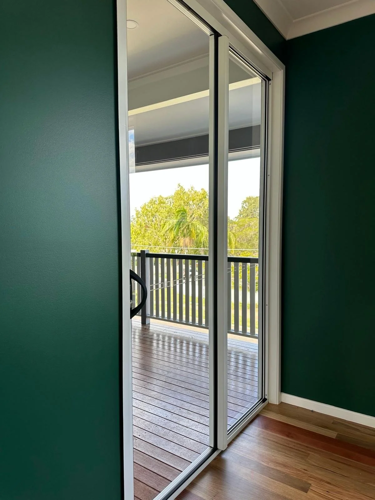 Open sliding glass door leading to a small balcony with black railing, view of green trees outside, green interior wall, and wooden floor.