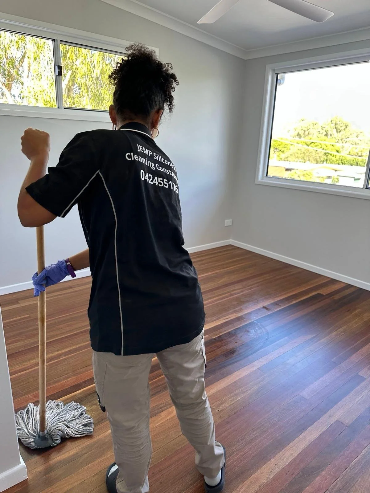 Person mopping wooden floor in a bright, empty room with large windows.