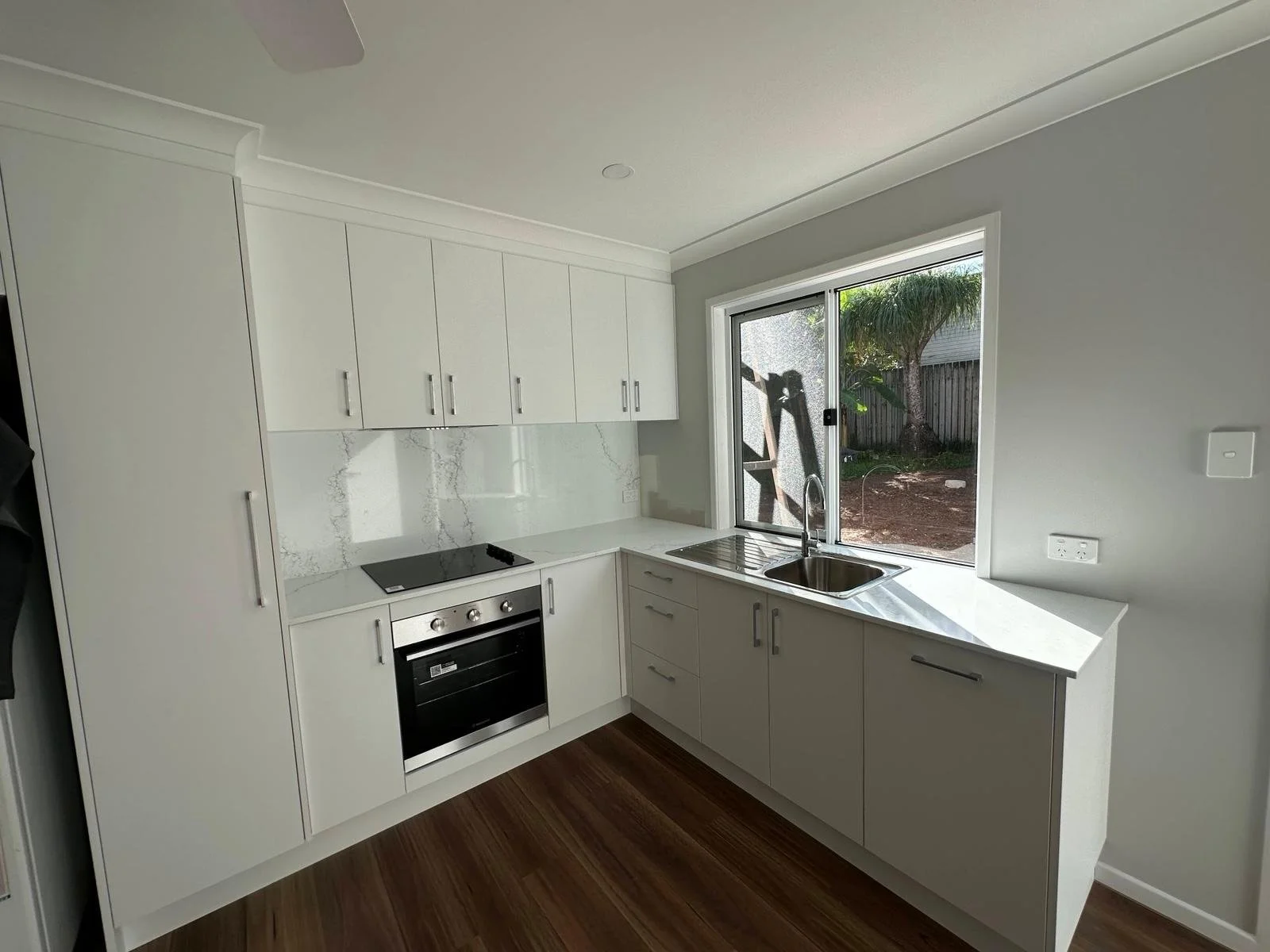 Modern kitchen with white cabinets, marble backsplash, black cooktop, stainless steel oven, stainless steel sink, and large window overlooking backyard with tree and wooden fence.