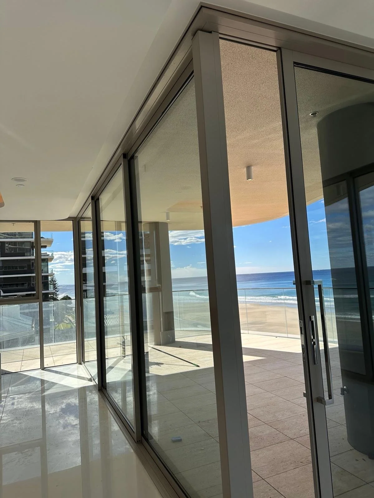 View of a balcony with glass sliding doors overlooking a beach and ocean under partly cloudy sky.