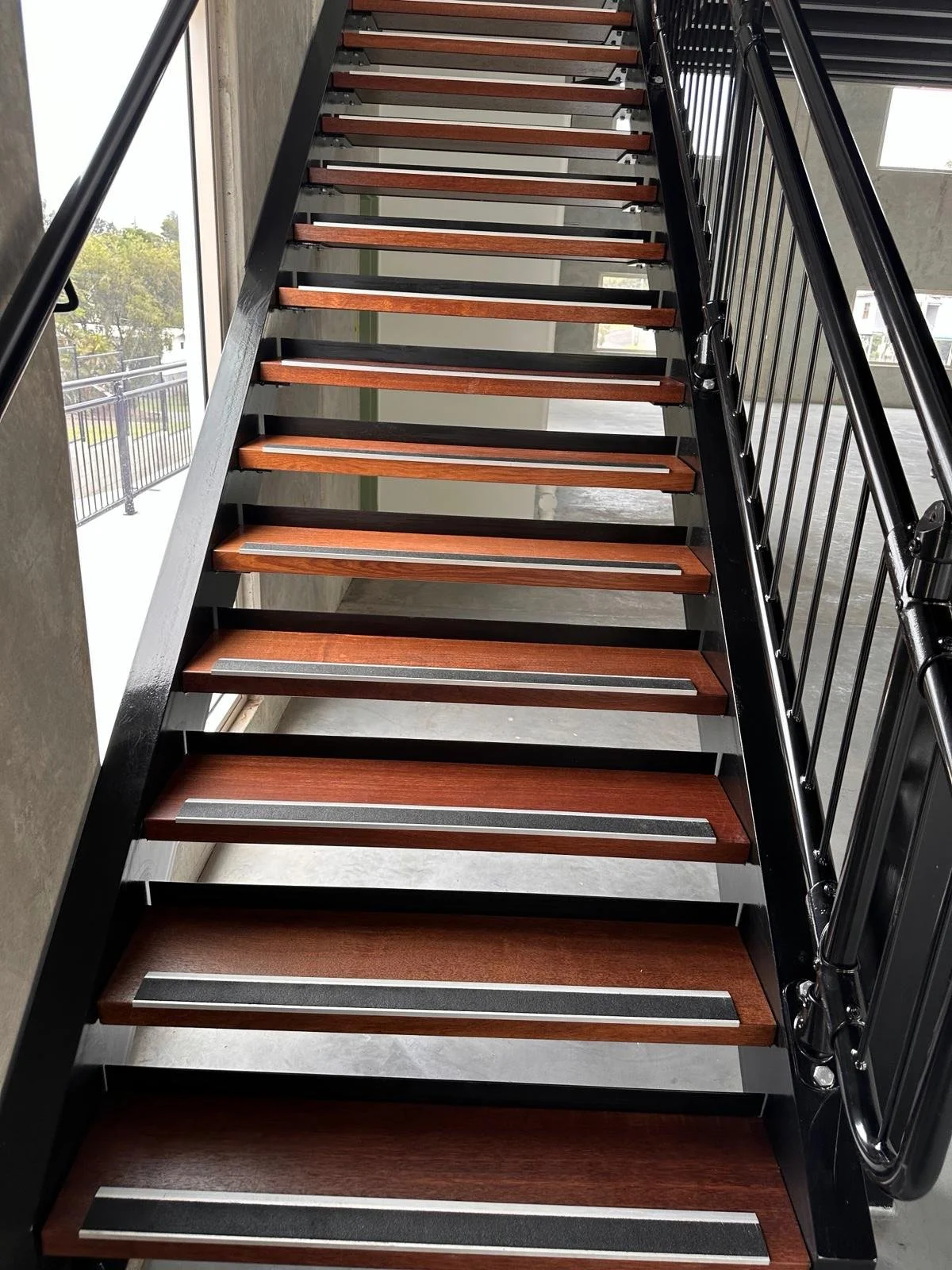 Interior view of a staircase with wooden treads, metal anti-slip strips, black railings, and large windows showing an outdoor balcony with a black railing and trees in the background.