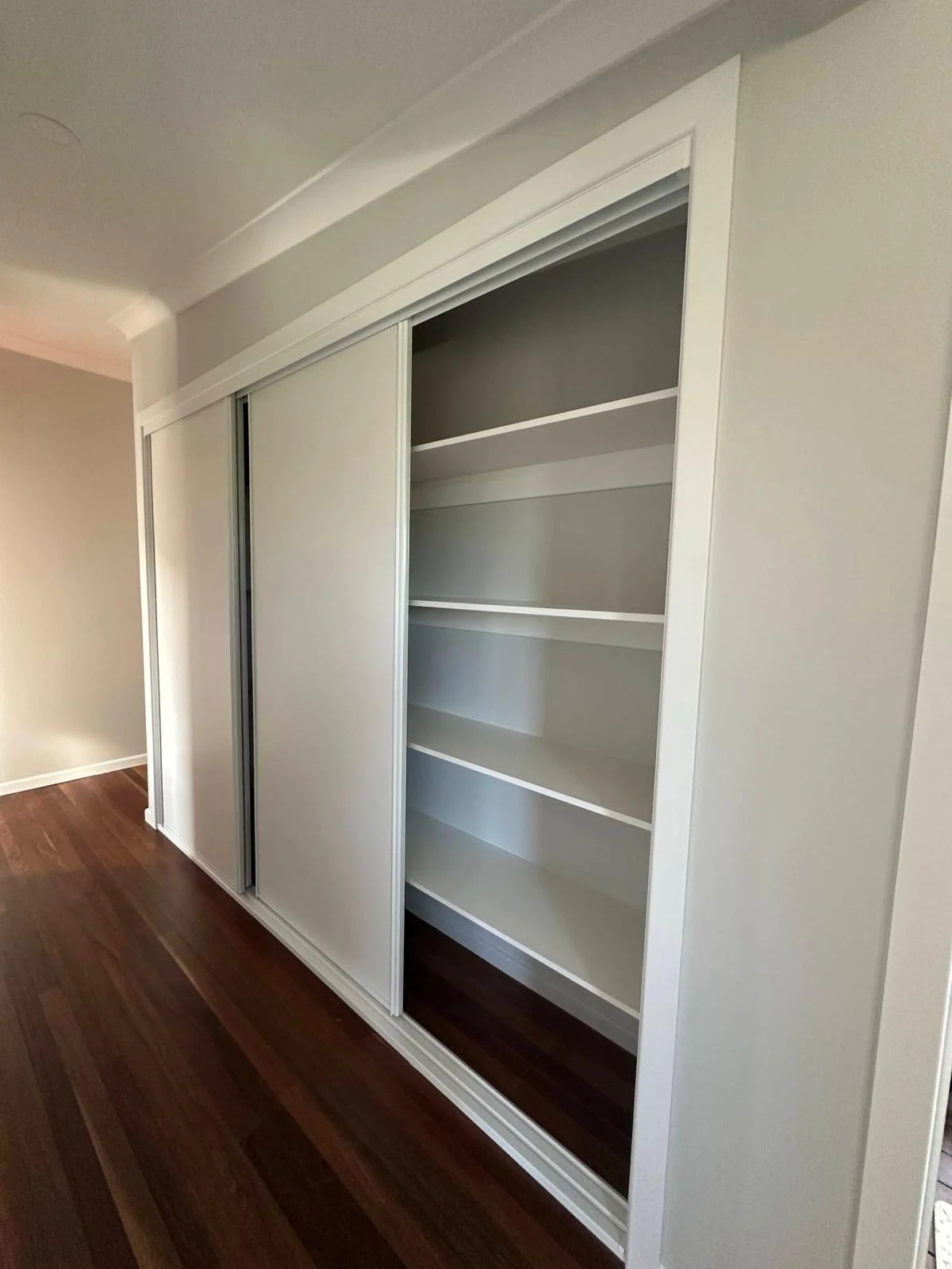 Empty built-in closet with sliding white doors, some open to reveal shelves inside, in a room with hardwood floors and light-colored walls.