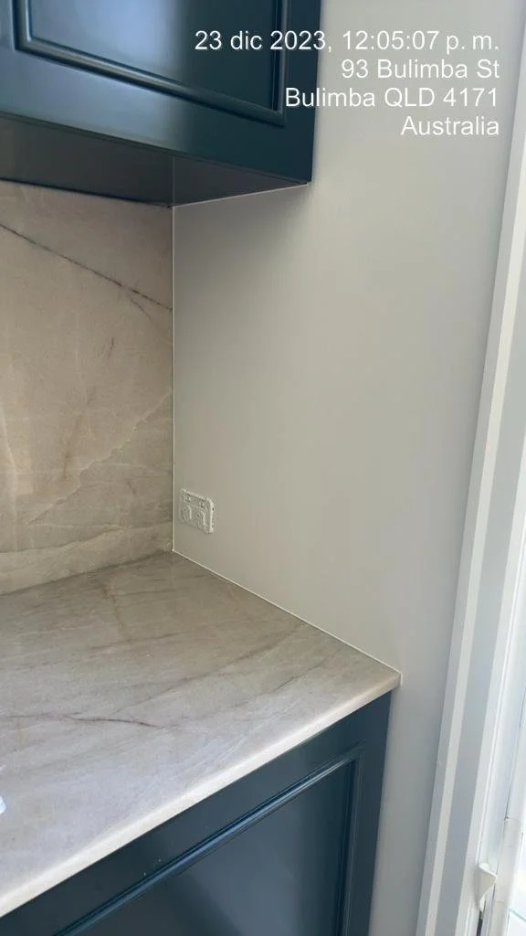 Close-up of a kitchen corner with beige stone countertop and backsplash, white wall, power outlet, and dark cabinets, with timestamp and address overlay