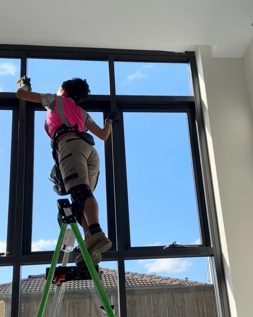 A person standing on a step ladder cleaning large window panes indoors during daytime, with a blue sky and some clouds visible outside.