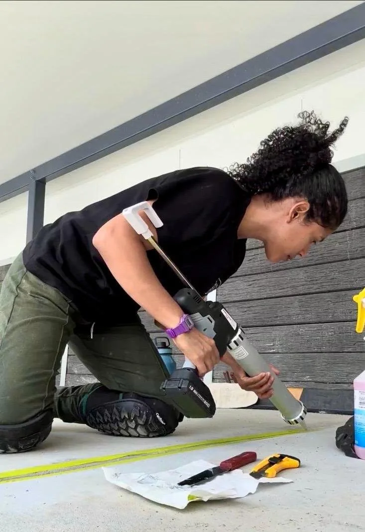 Woman kneeling on the floor, using a caulking gun with tools and supplies nearby.