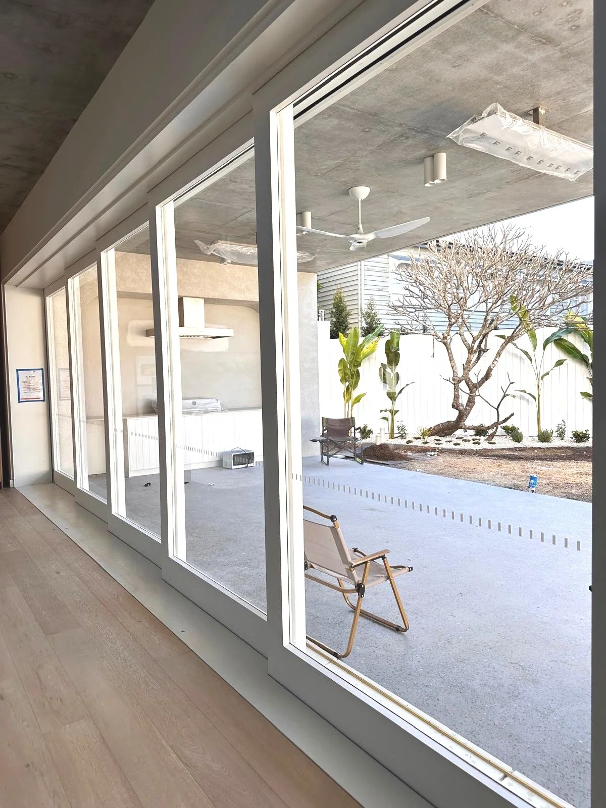 View of a backyard patio area through large glass sliding doors, featuring a wooden lawn chair, a tree with no leaves, some green plants, outdoor ceiling fan, and a hose on a landscaped ground.