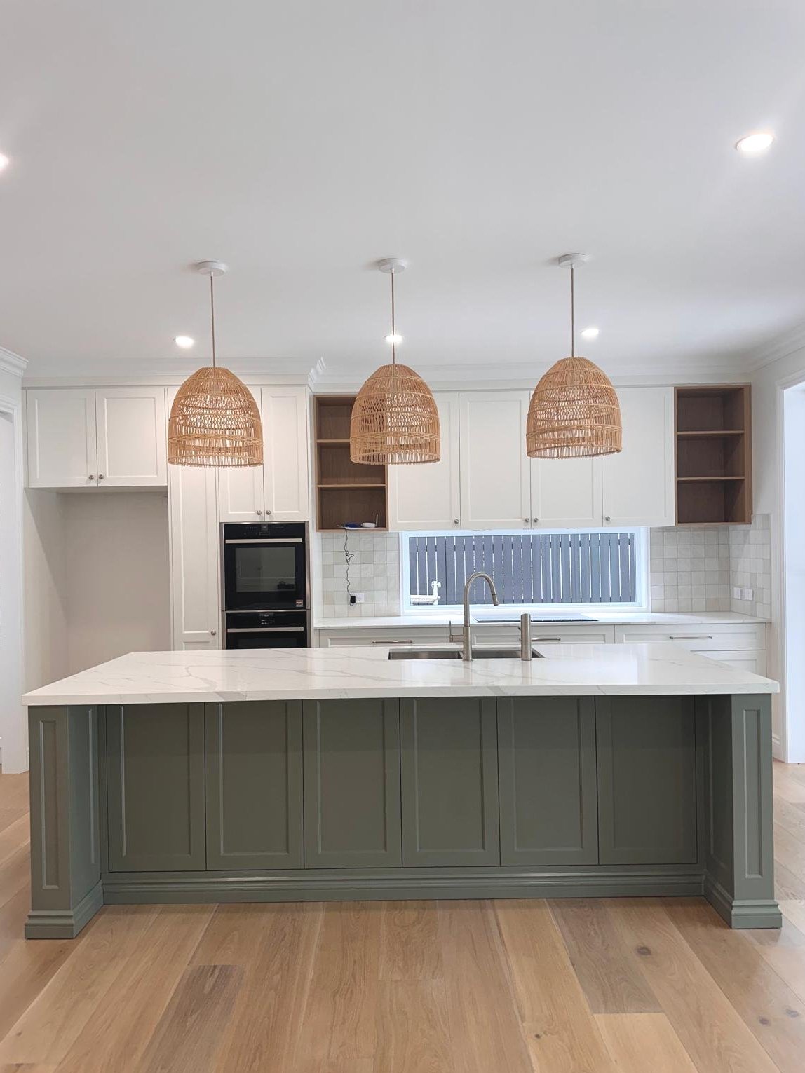 Modern kitchen with white cabinets, a large island with a green base and white marble countertop, three woven pendant lights, and a window above the sink.