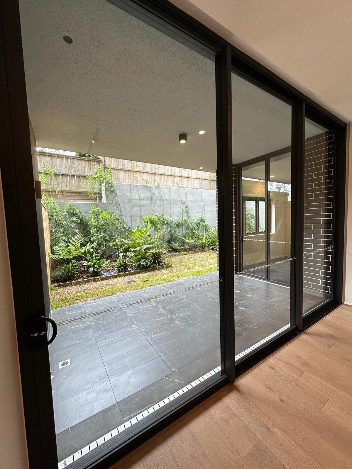 View of a small backyard garden through a large glass sliding door with black frames, showing lush green plants, a gray concrete wall, and a wooden fence; inside has wooden flooring and a beige ceiling with recessed lighting.