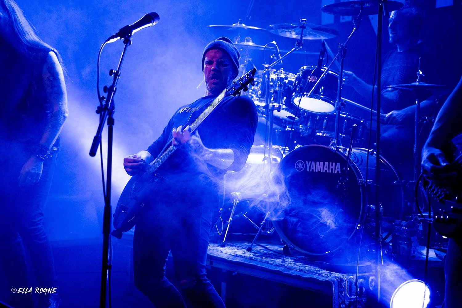 Musiker spiller gitar på scenen med blå belysning og damp i luften, med trommesett i bakgrunnen.