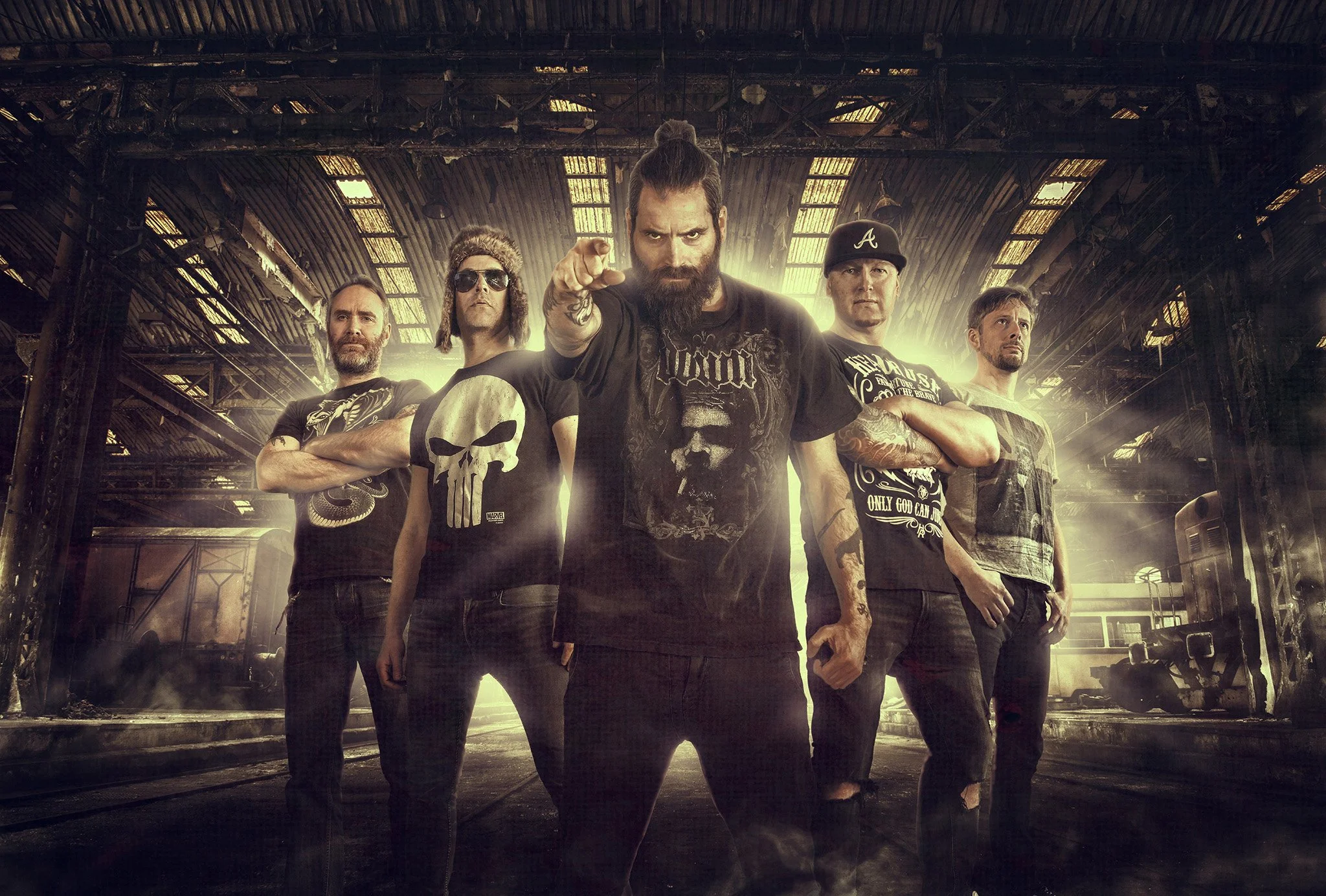 En gruppe fem menn står i en industriell miljø med rustne metallbjelker og et gammelt tog i bakgrunnen, med lys som skinner gjennom taket bak dem.