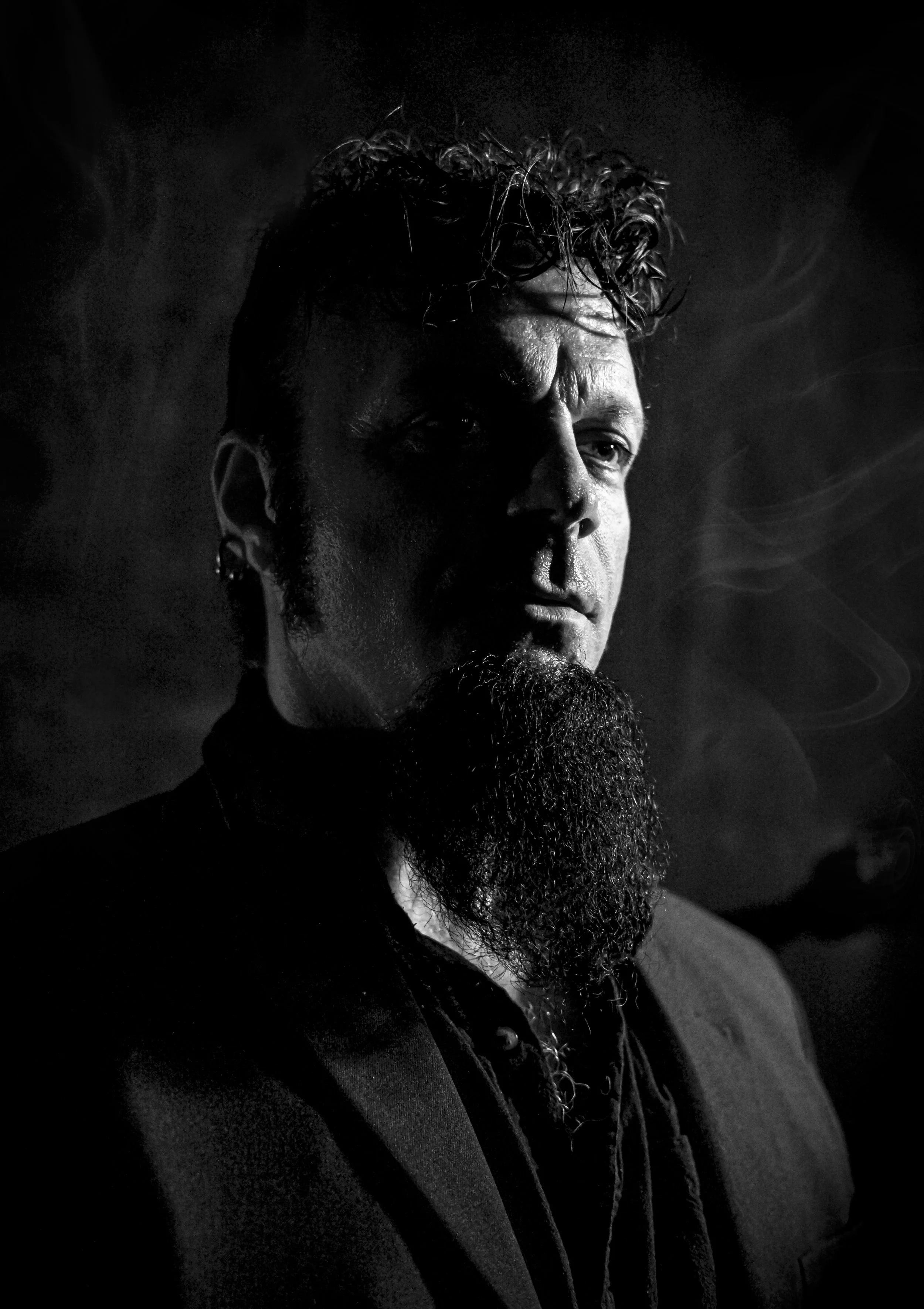 Sort-hvitt bilde av en mann med skjegg, som røyker og er kledd i mørk jakke, med røyk rundt ham, i lavt lys.