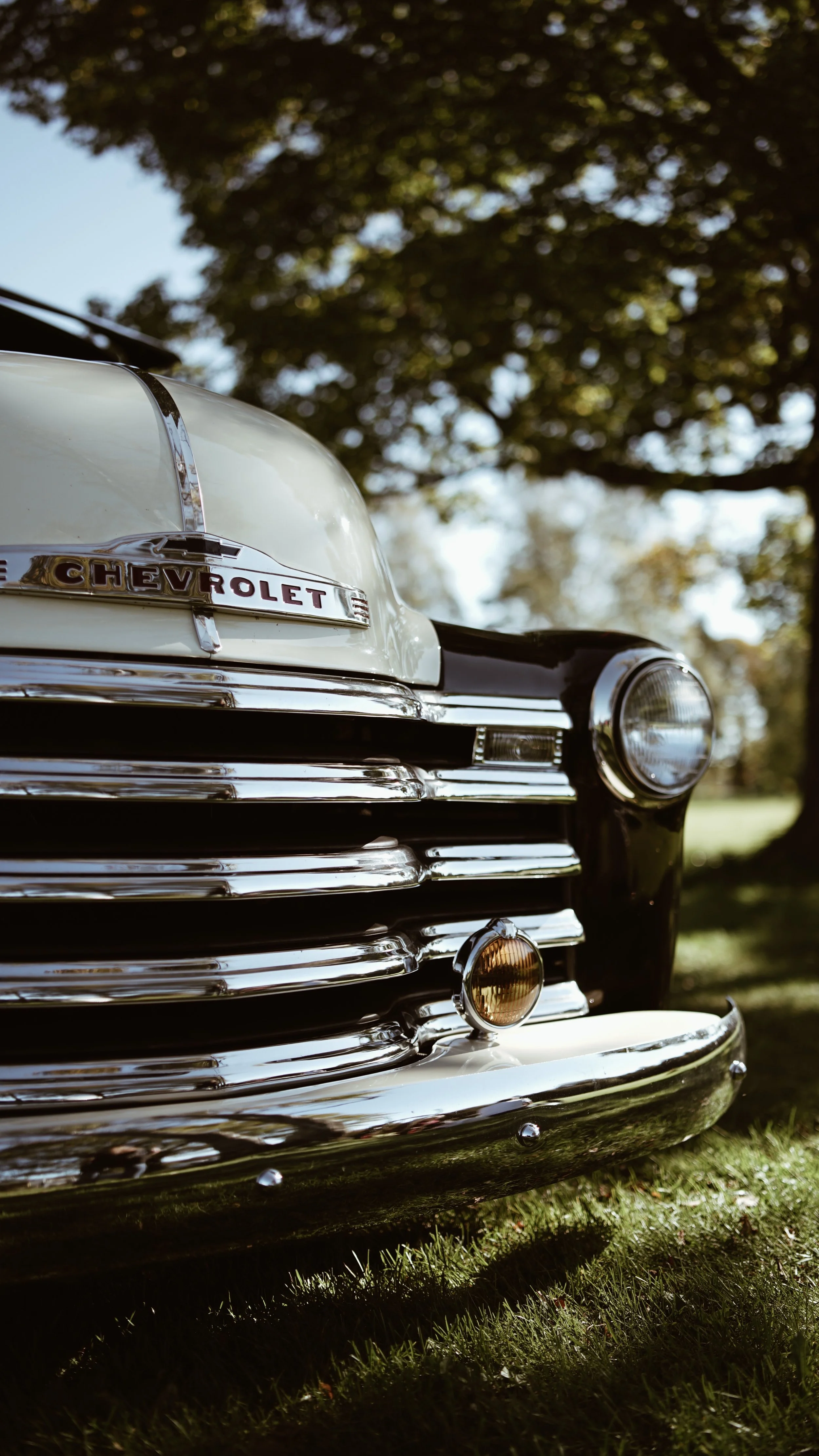 Nærbilde av fronten på en klassisk Chevrolet-bil med kromdetaljer og et Chevrolet-emblem, parkert under trær.