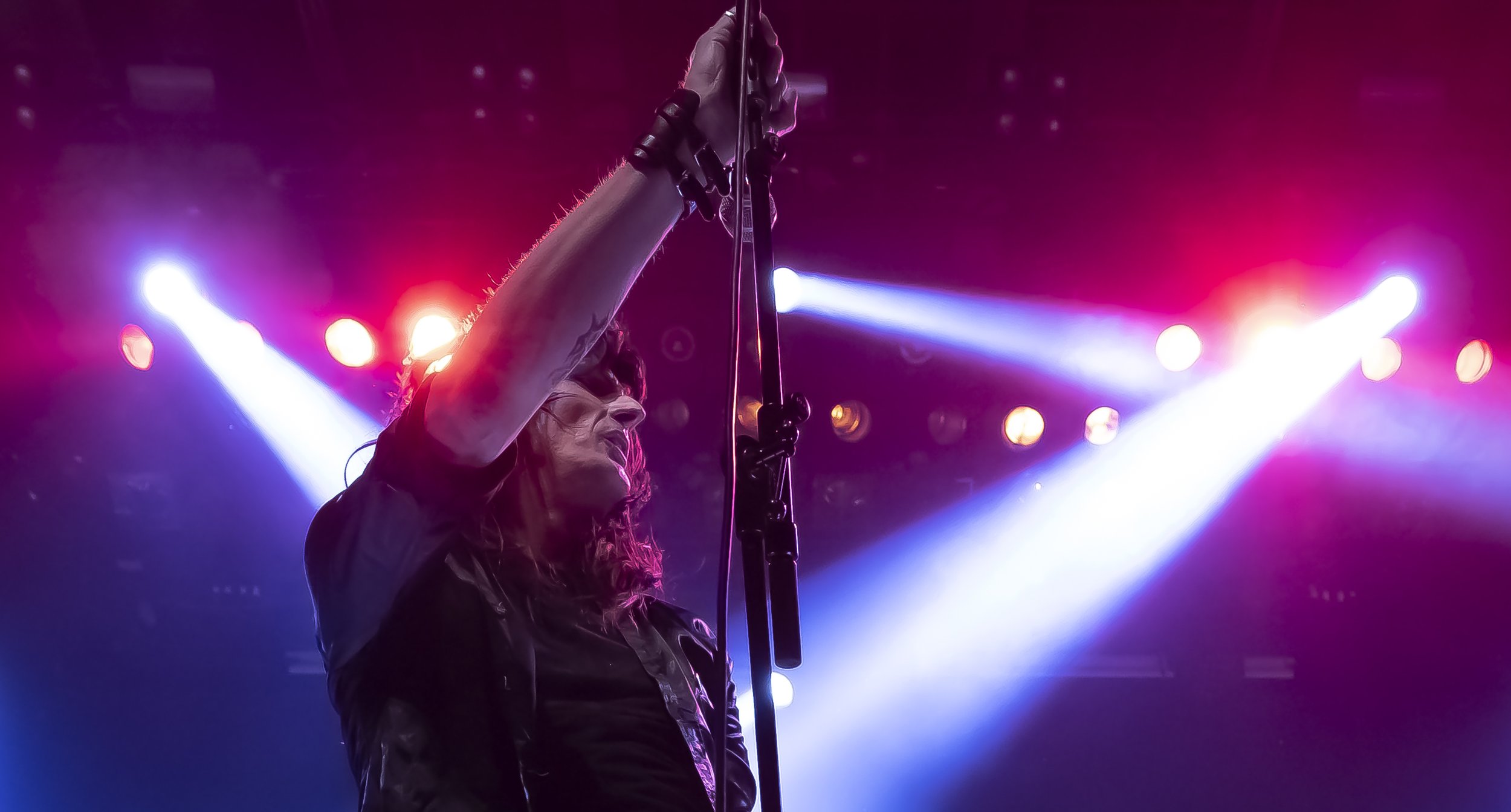 En person med langt, krøllet hår som opptrer på en konsert, med lyse fargerike scenelys i bakgrunnen.