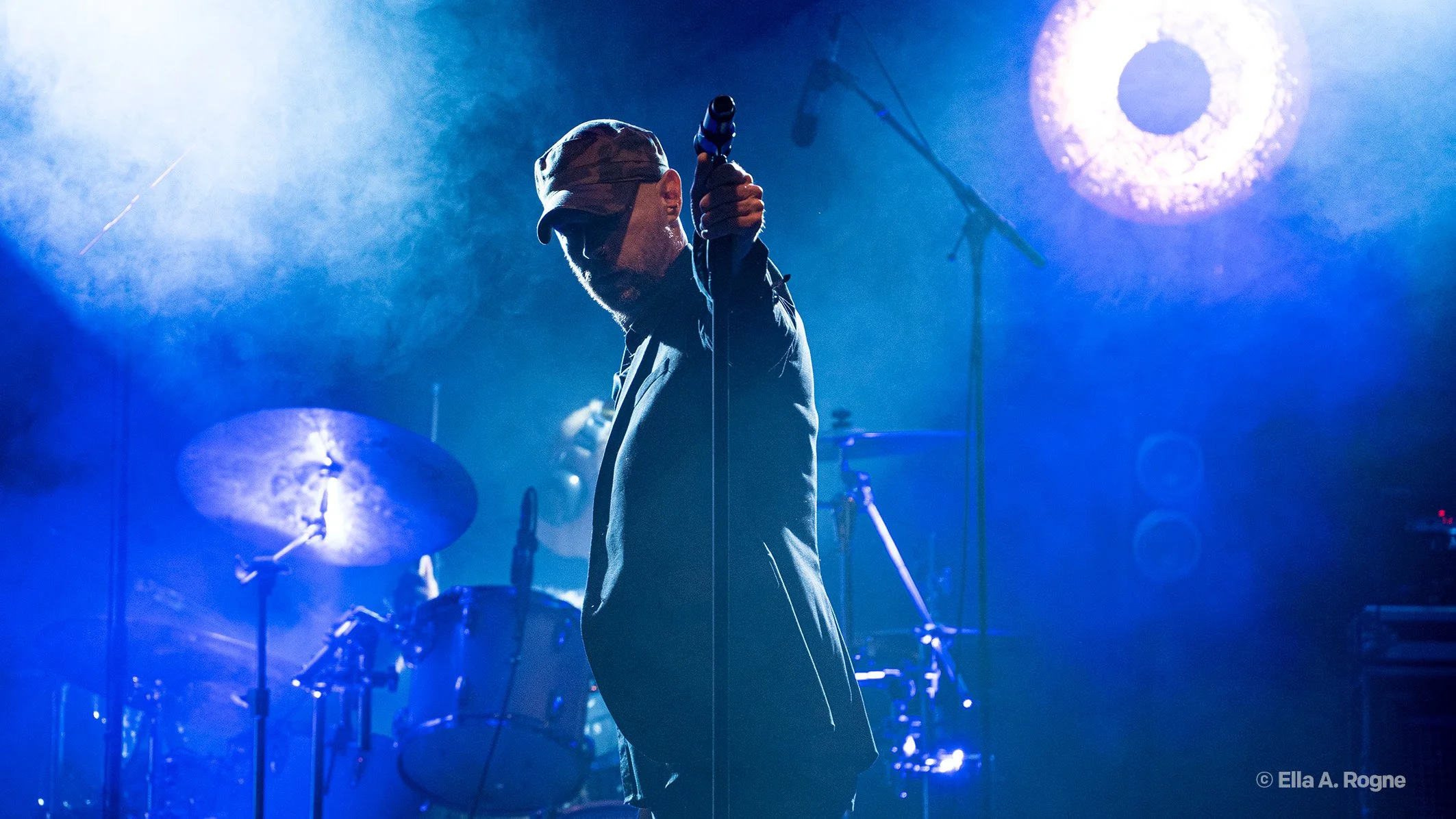 En mann på scenen med mikrofon, omgitt av blått lys og tåke, med trommer i bakgrunnen.
