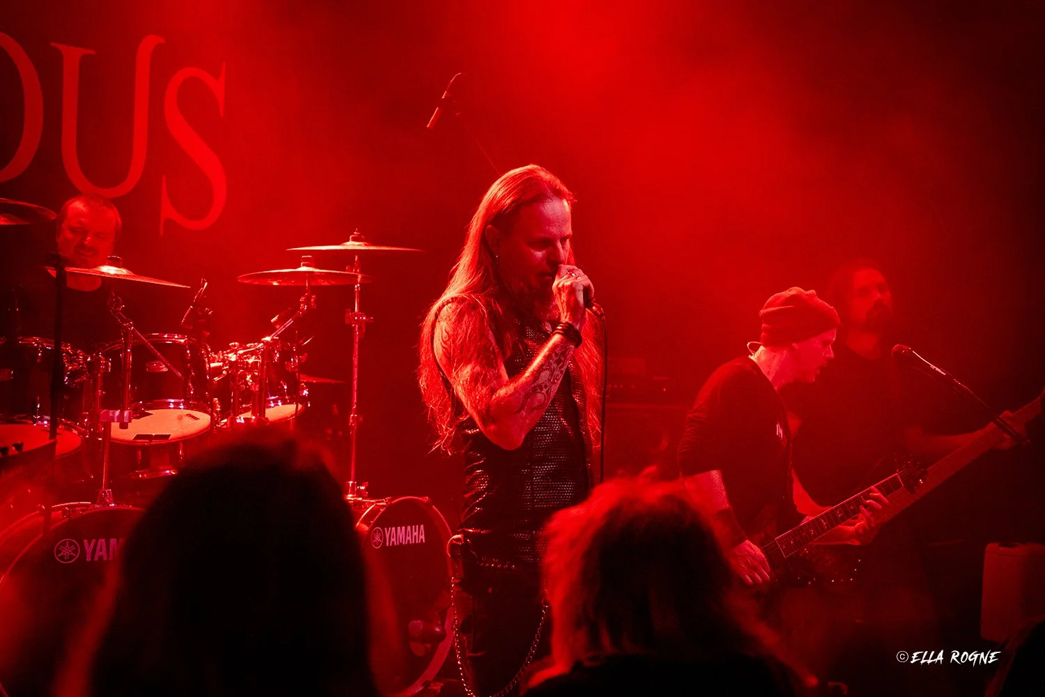 En rockeband som spiller på en konsert med rød belysning, med et kvinnelig vokal- og gitarist i sentrum, og en trommeslager og en bassgitarist i bakgrunnen.