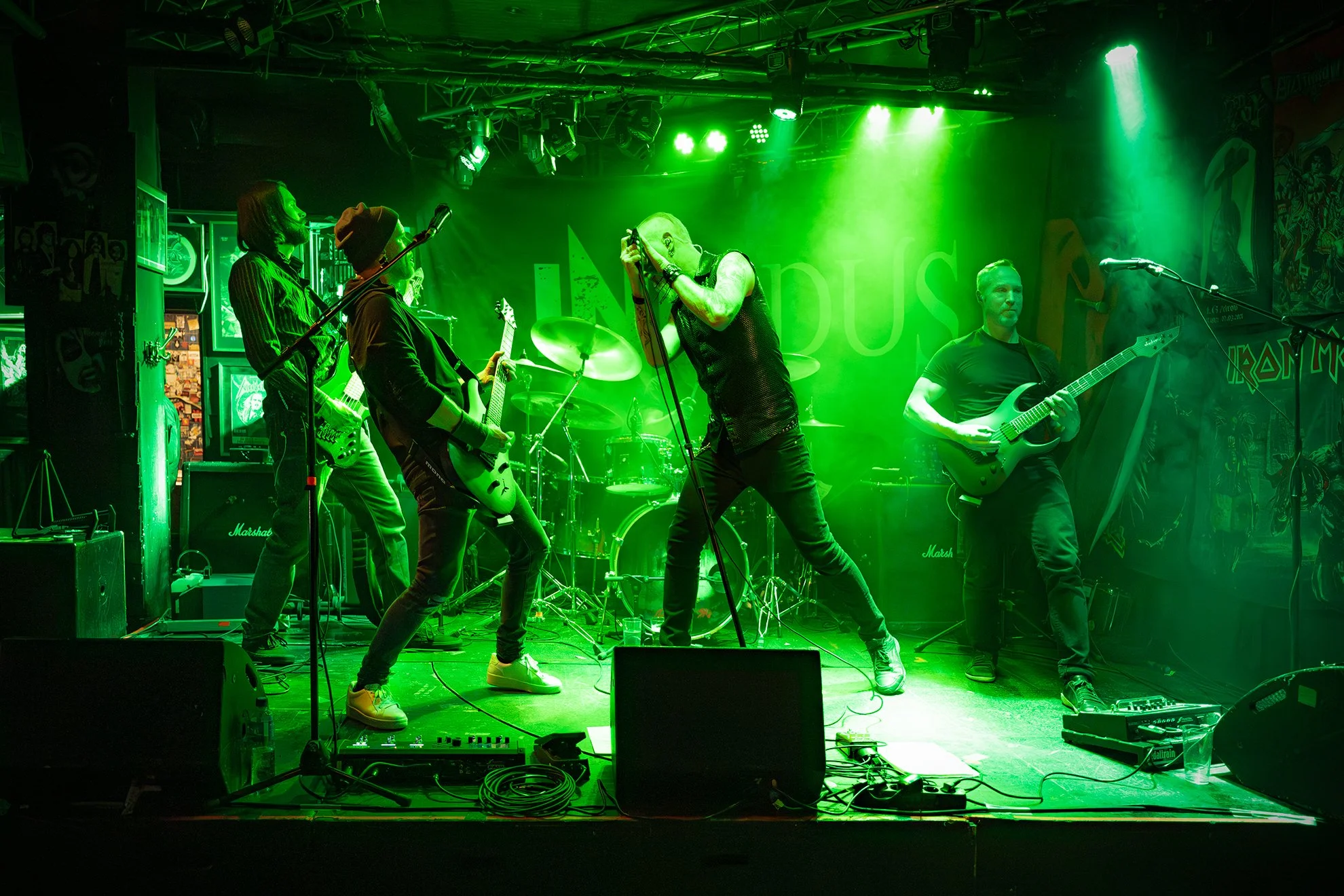 En livemusikkonsert med fem musikere på scene, omgitt av grønt lys. Musikerne spiller gitarer, og det er en trommesett i bakgrunnen. Det er også forsterkere og annet utstyr på scenen.