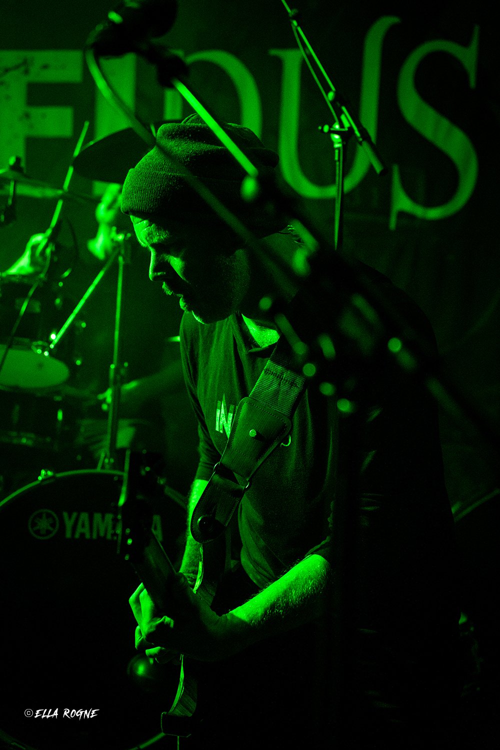 En person spiller gitar på en scene med grøn belysning, bakgrunn med store grønne bokstaver.