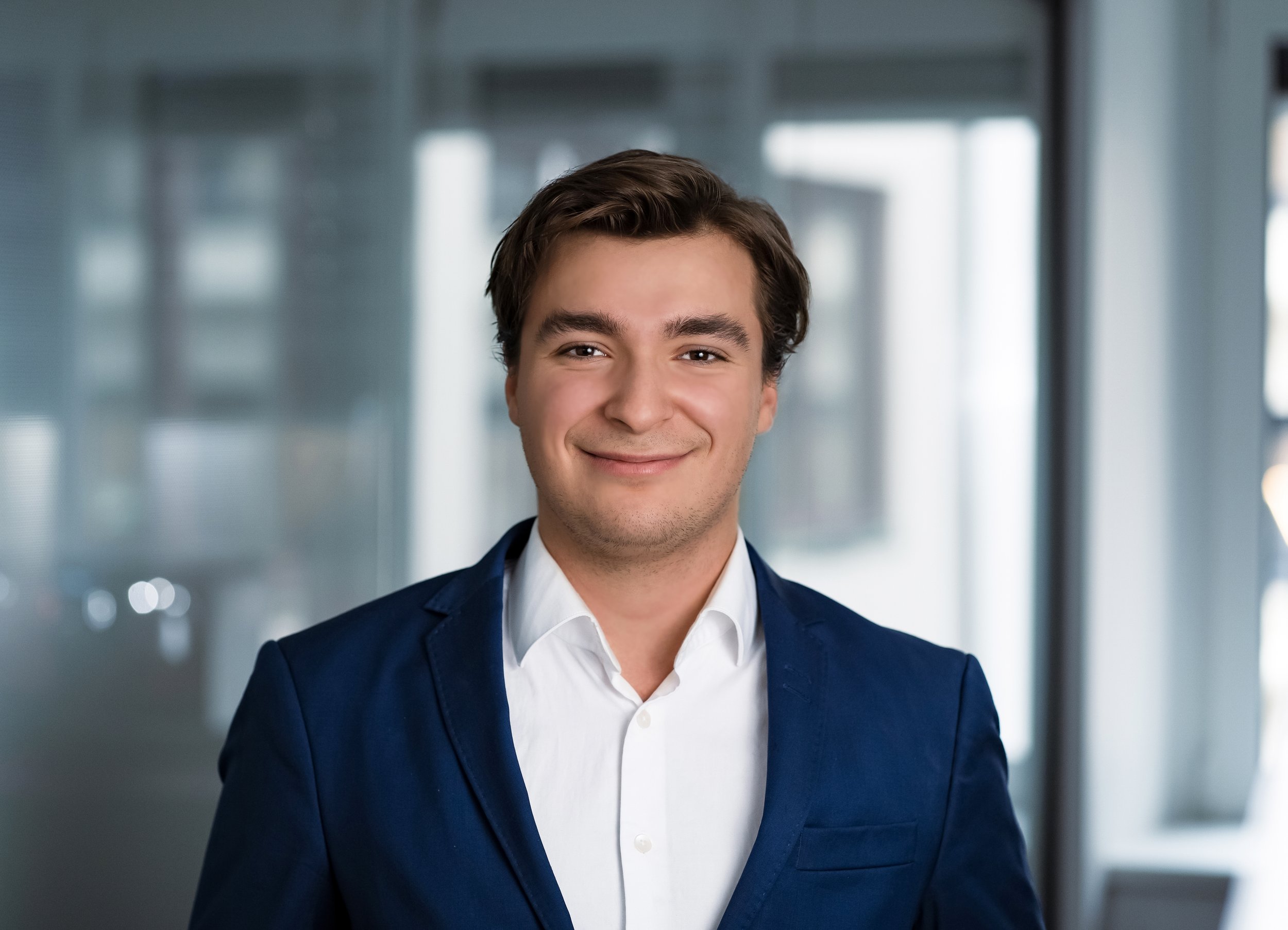 En ung mann iført en hvit skjorte og en blå blazer smiler i en kontorbygning.