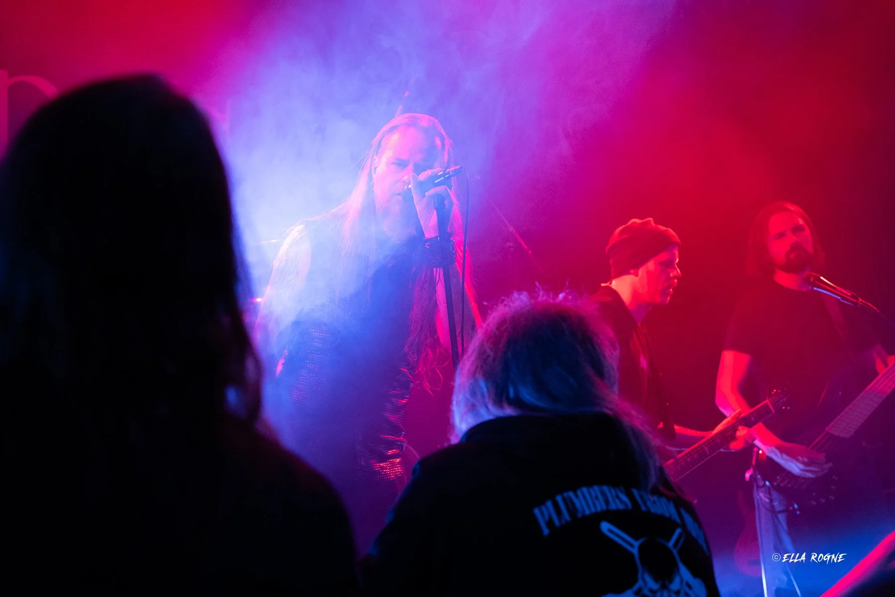 Musikere på scenen med farget lys, en kvinne synger inn i en mikrofon, mens to menn spiller gitar i bakgrunnen, foran publikum.