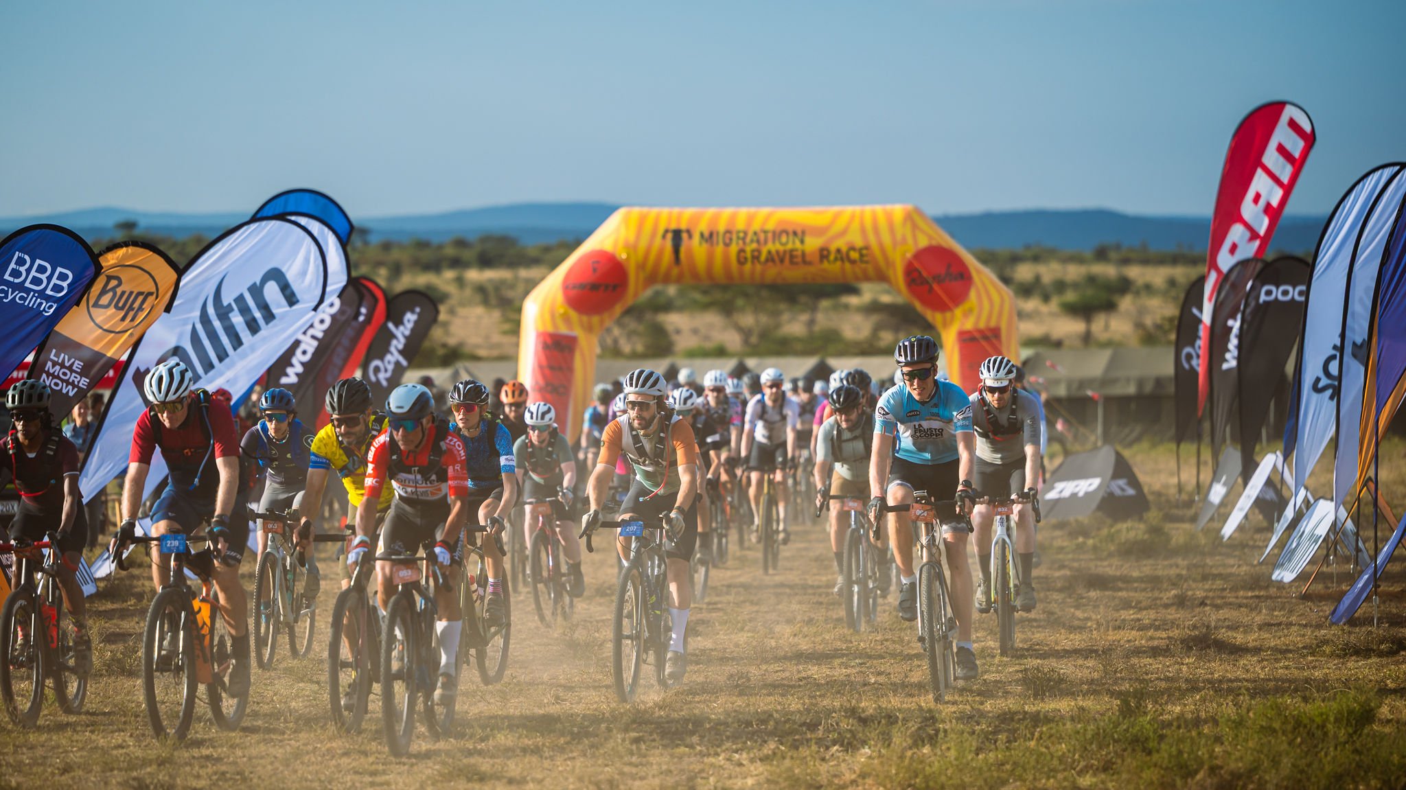 A large group of cyclists participating in a gravel race, riding on a dirt path through a field with banners on either side and a large yellow inflatable arch reading "Migration Gravel Race" in the background, under a partly cloudy sky.
