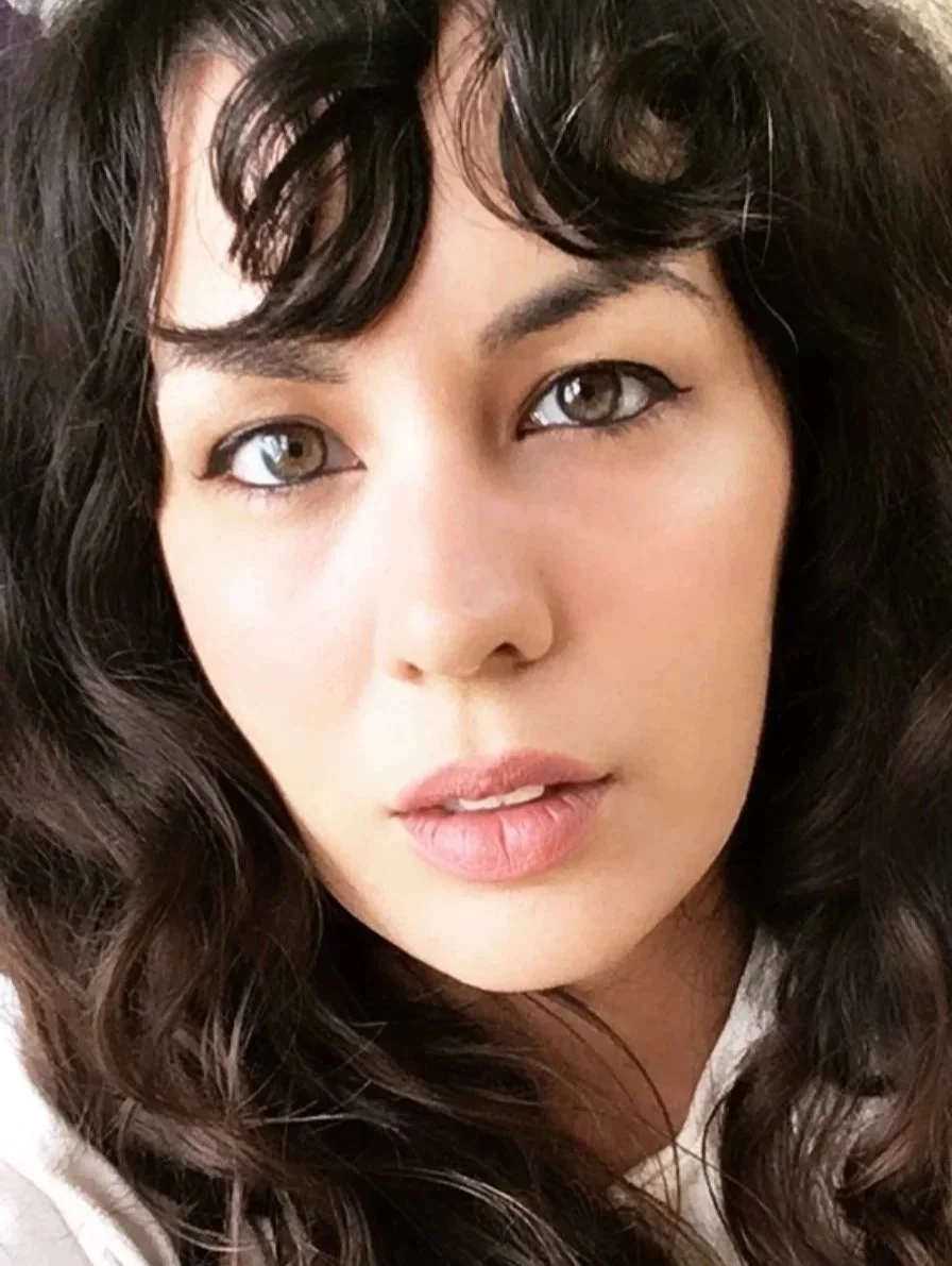 Close-up of a woman with dark wavy hair, light skin, and hazel eyes, looking directly at the camera.